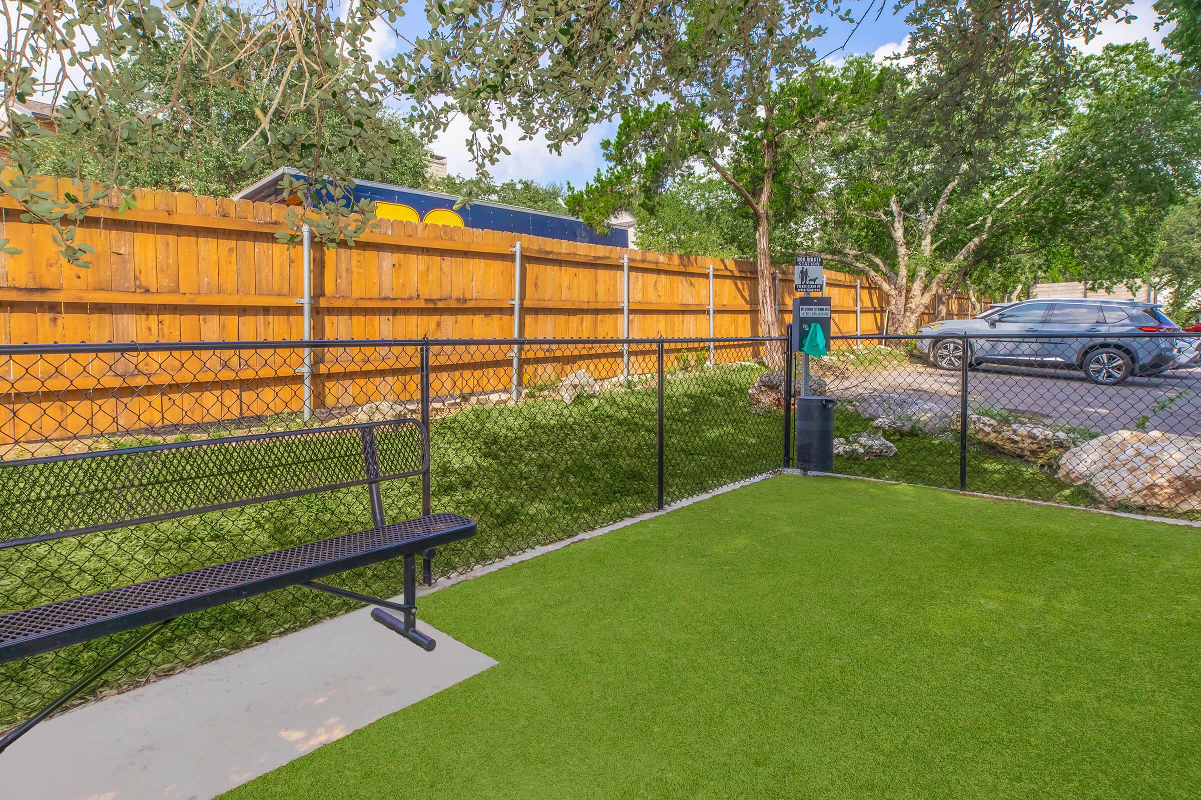 A small fenced area with artificial grass, featuring a bench on the left. Behind it, a wooden fence provides privacy. A car is parked nearby, and there are some rocks and trees in the background, creating a natural setting.