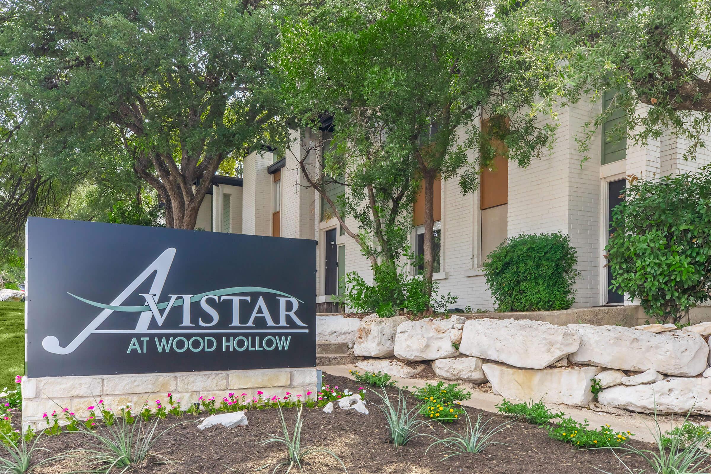 Sign for "Avistar at Wood Hollow" displayed in front of a landscaped area with green foliage and flowers, next to a stone border. The building behind features a modern architectural design with multiple windows and shades.
