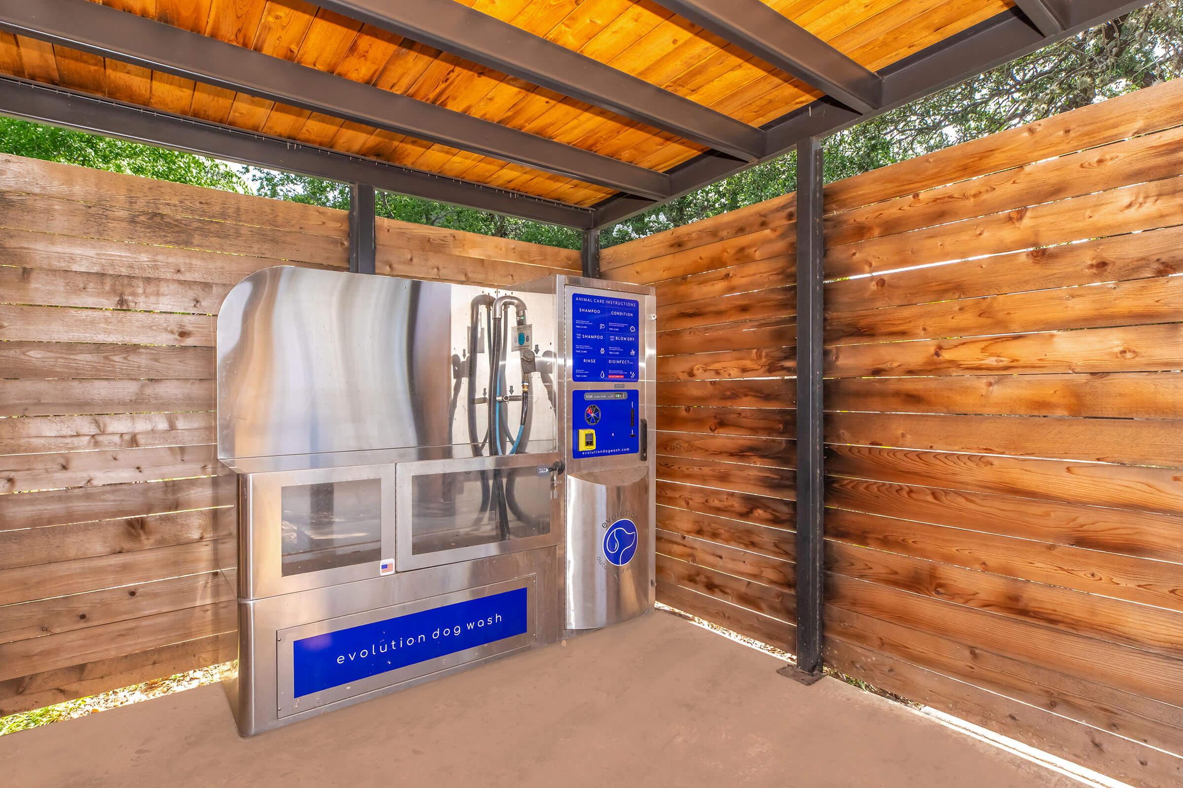 A stainless steel dog washing station situated under a wooden shelter, featuring a control panel with instructions and options for washing pets. The surrounding walls are made of wooden slats, creating a rustic outdoor space for dog grooming.