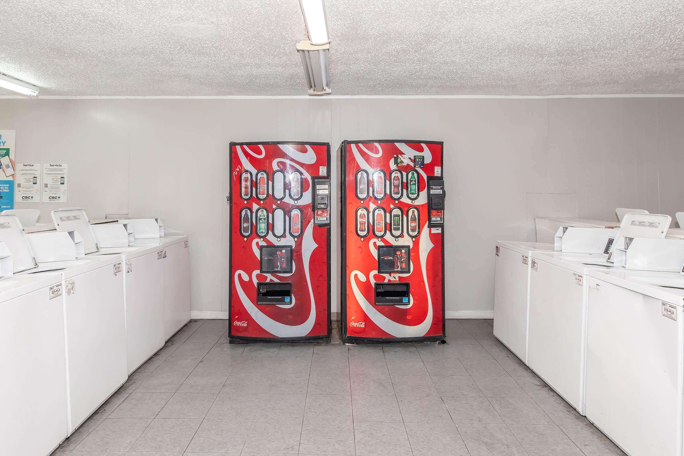 A laundromat interior featuring rows of white washing machines and two red Coca-Cola vending machines. The space has a gray tiled floor and fluorescent lighting, creating a bright and clean atmosphere.