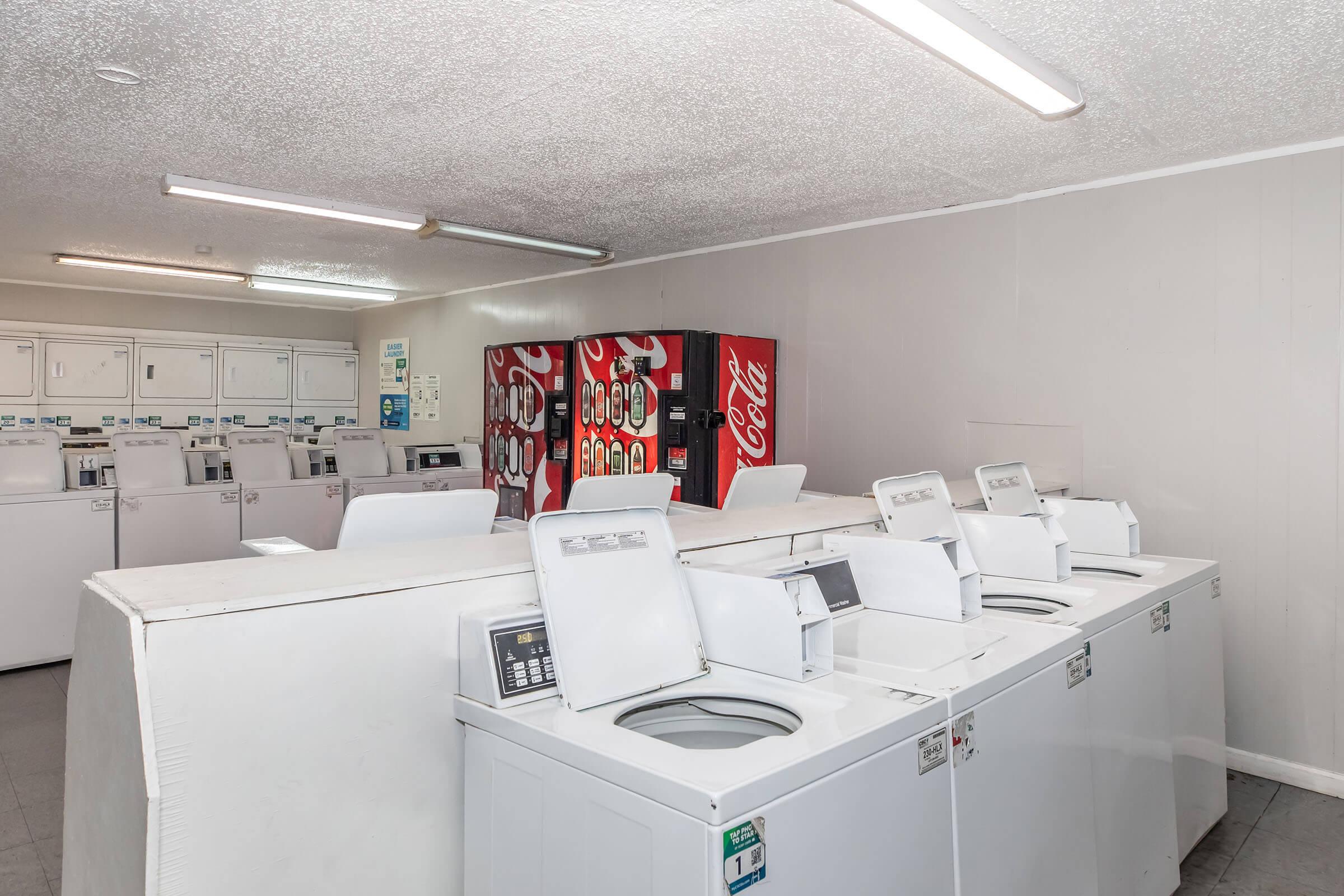 A clean, well-lit laundry room with several white washing machines lined up against a wall. In the background, there is a vending machine displaying Coca-Cola products. The space appears organized and inviting, suitable for doing laundry.