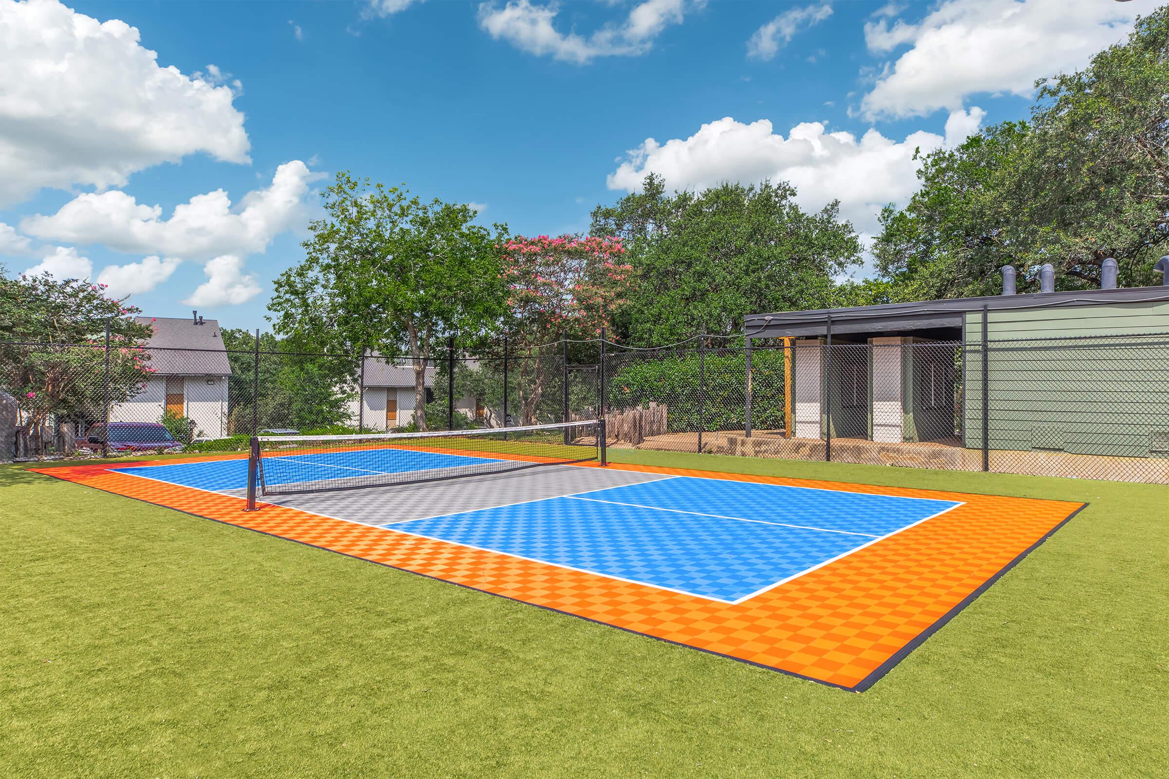 A vibrant tennis court with blue and orange markings, surrounded by greenery. The court features a net in the center and is set against a backdrop of residential buildings and trees. The sky is partly cloudy, creating a bright and inviting atmosphere.