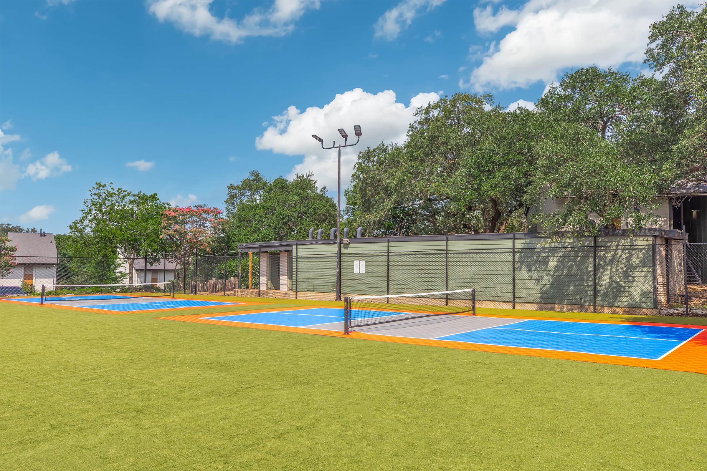 A bright outdoor recreational area featuring two multi-colored tennis courts with blue and orange surfaces, surrounded by green grass. In the background, there are trees and a building, with a clear blue sky and scattered clouds above.