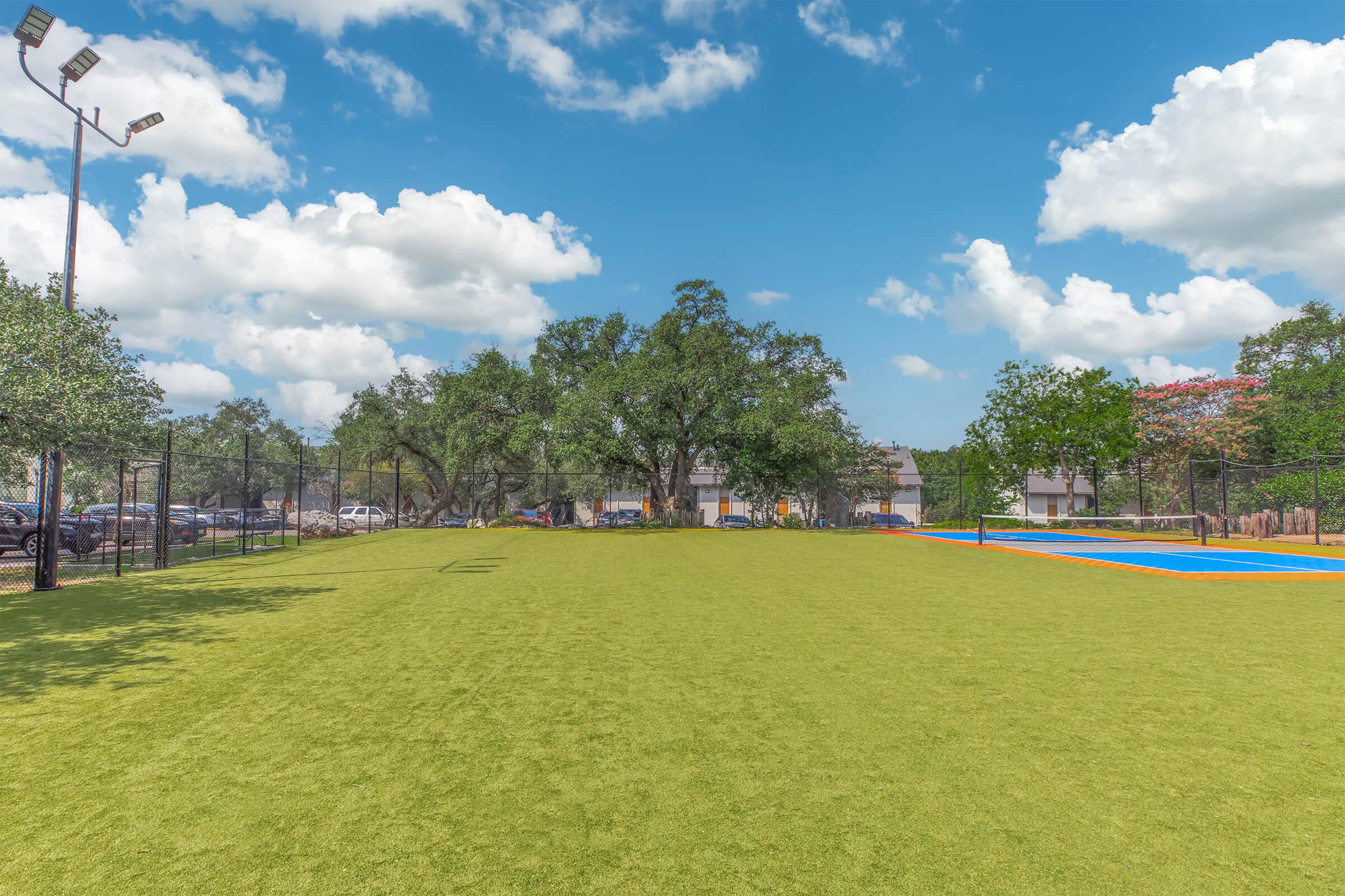 A wide view of a park featuring a well-maintained artificial grass area under a blue sky with scattered clouds. In the background, there are several trees and a residential building. Two basketball courts can be seen on the right, highlighting vibrant colors. The setting is bright and inviting, ideal for outdoor activities.
