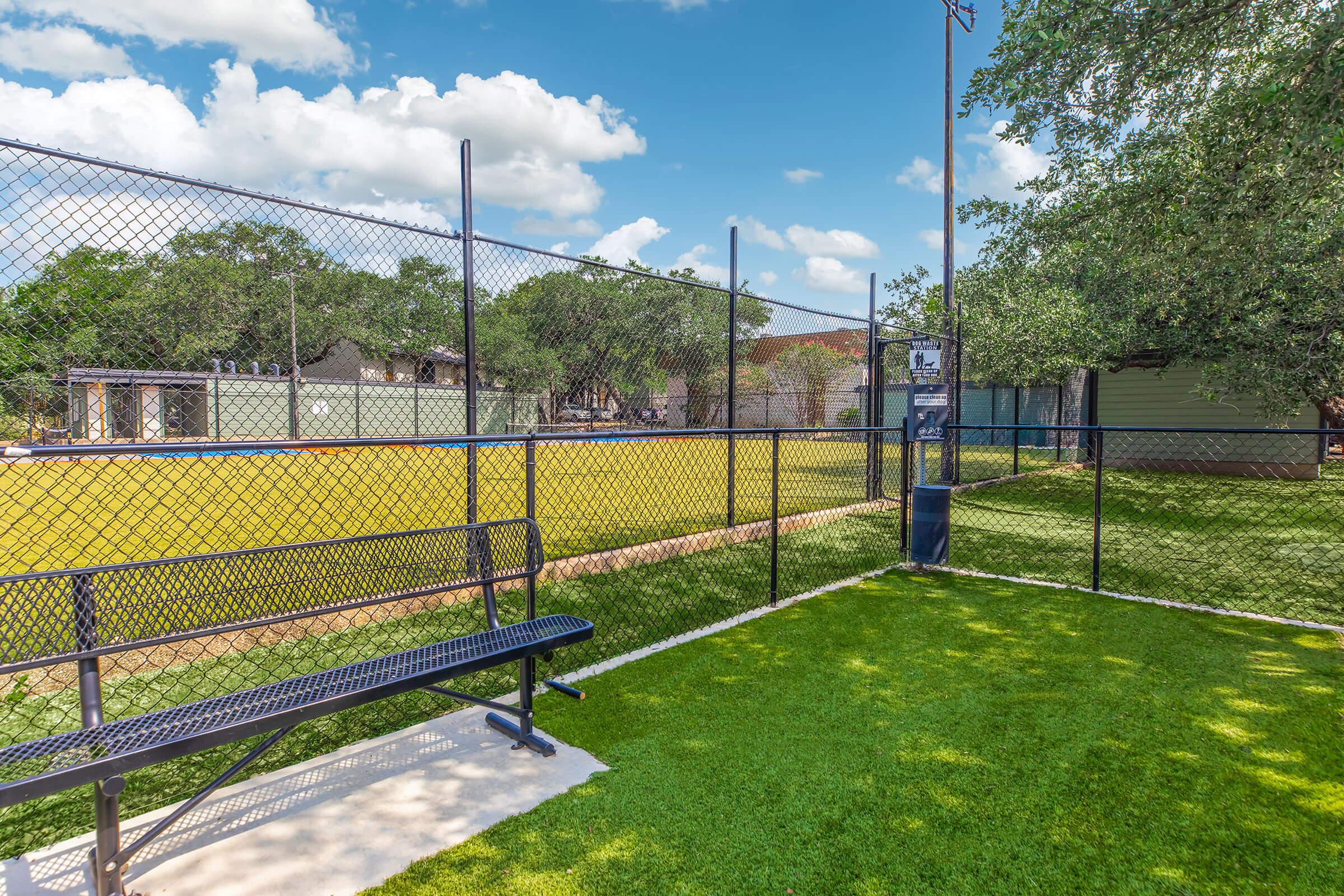 A well-maintained sports field enclosed by a chain-link fence, featuring a grassy area and a bench for spectators. There are trees in the background, and the sky is blue with a few scattered clouds. The field appears ready for play, indicating an inviting outdoor space for sports activities.