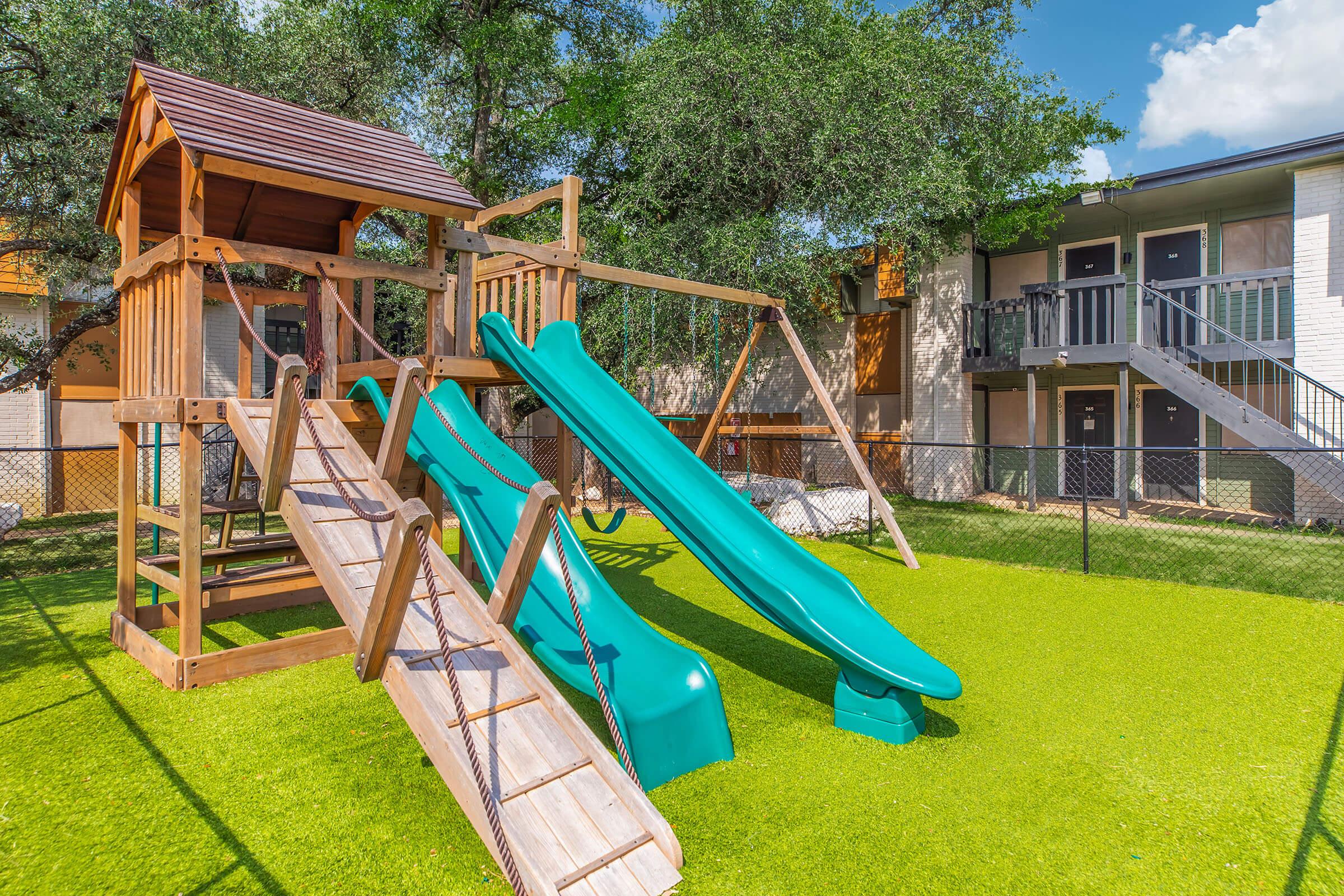 A wooden playground structure featuring two green slides, a climbing ramp, and swings. The playground is set on bright green artificial grass, surrounded by trees and adjacent to an apartment building with balconies. The scene is well-lit, showcasing a clear blue sky.