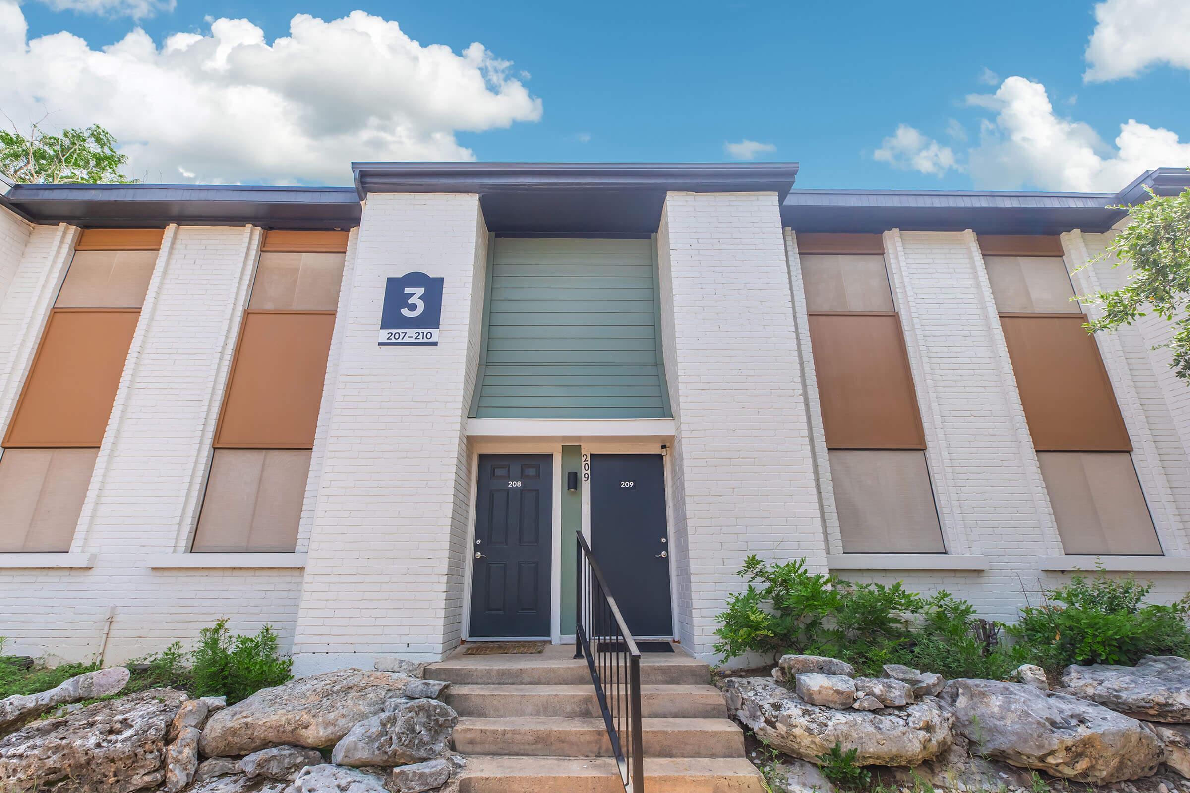 A two-story building featuring a white brick facade with large windows partially covered by shades. The entrance has a set of stairs leading to two black doors. A blue panel indicates apartment number 3, located at 207-210. Surrounding greenery and a bright blue sky with clouds enhance the setting.