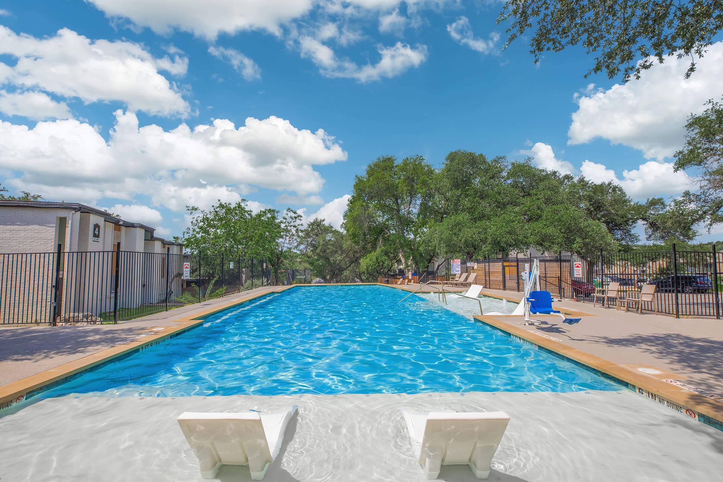 A clear blue swimming pool surrounded by a spacious deck, featuring white lounge chairs and a fence. Lush green trees and a bright blue sky with fluffy white clouds are visible in the background, creating a relaxing outdoor atmosphere.