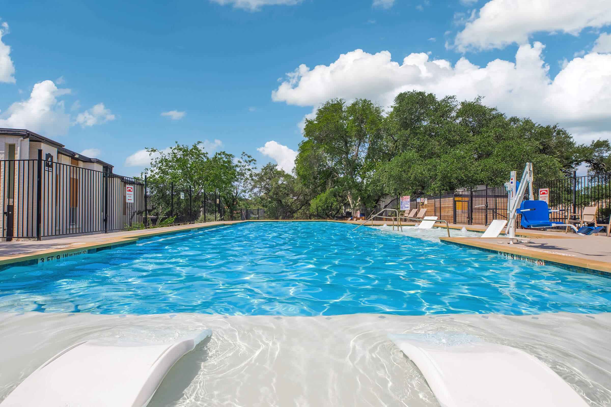 A clear blue swimming pool surrounded by a fence, lounge chairs, and a few trees. The sky is bright with fluffy white clouds, creating a relaxing atmosphere. The pool features a wide ledge and a smooth surface reflecting the sunlight.
