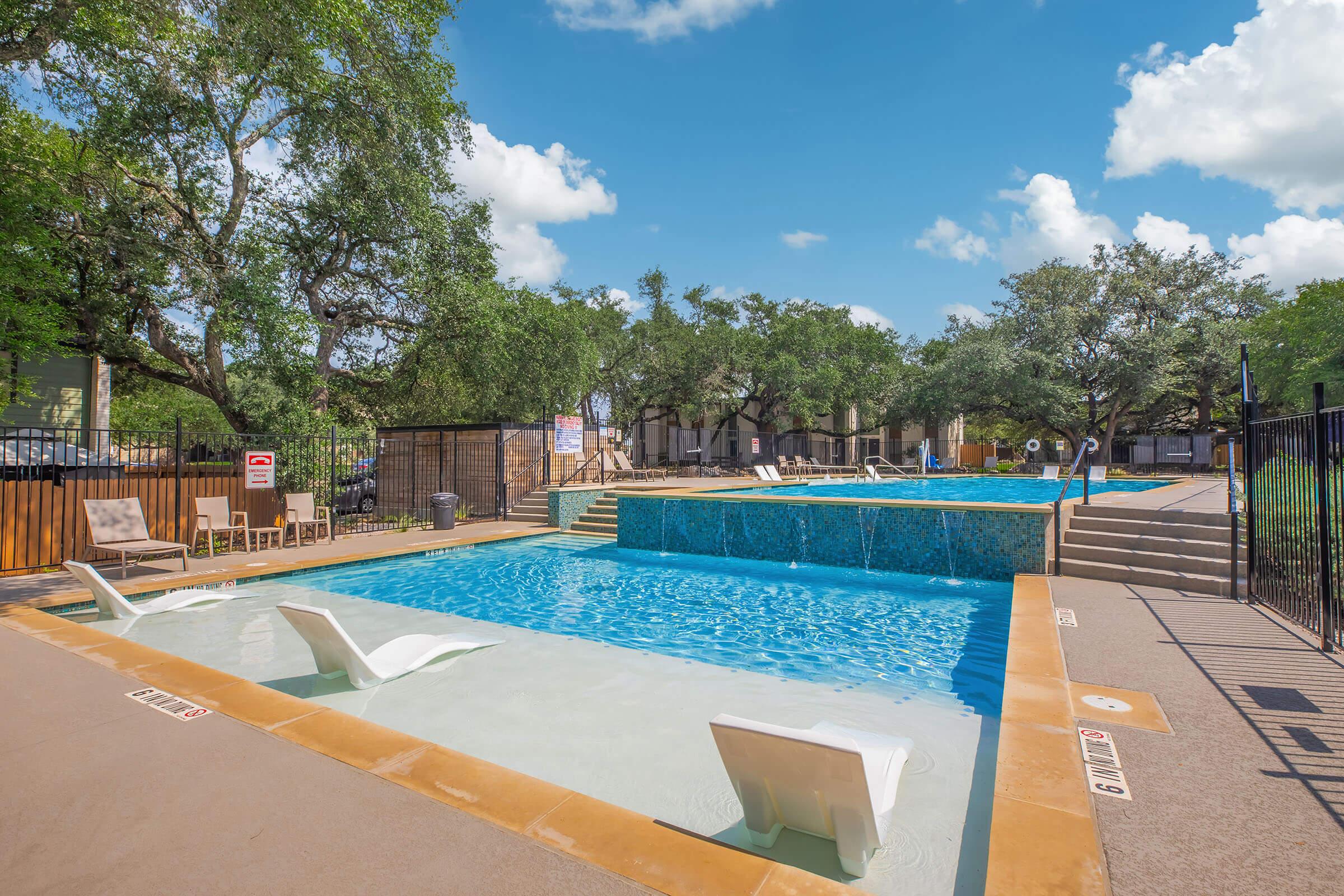 A scenic view of a swimming pool area featuring multiple pools, surrounded by trees and a clear blue sky. There are poolside chairs and a gated fence. The pool surfaces reflect sunlight, and the area is well-maintained, creating a relaxing atmosphere for visitors.