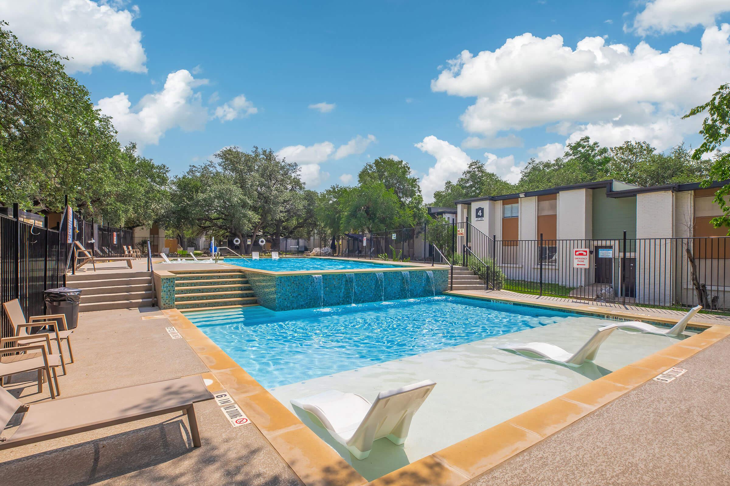 A scenic outdoor pool area featuring two pools with water features. One pool has submerged lounge chairs. Surrounding the pools are trees and a fence, with residential buildings in the background. The sky is bright blue with fluffy white clouds.