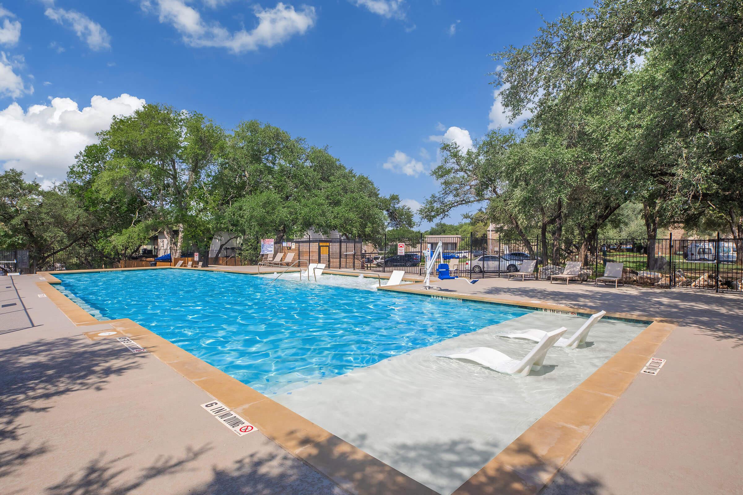 A clear swimming pool surrounded by a concrete deck, with several lounge chairs positioned around it. Lush green trees and a blue sky with fluffy clouds in the background. A fence encloses the pool area, and there are signs visible nearby.
