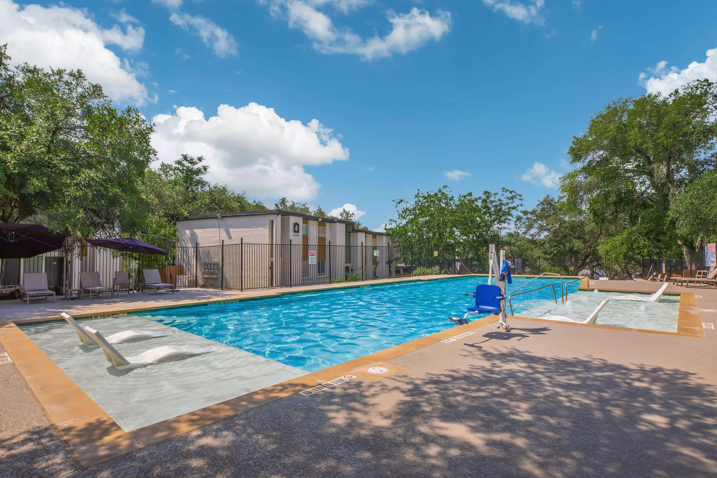 A sunny outdoor swimming pool surrounded by greenery. The pool features a bright blue water, a ladder for entry, and lounge chairs placed nearby. Shadows of trees and a clear blue sky with scattered white clouds enhance the inviting atmosphere of the leisure area.