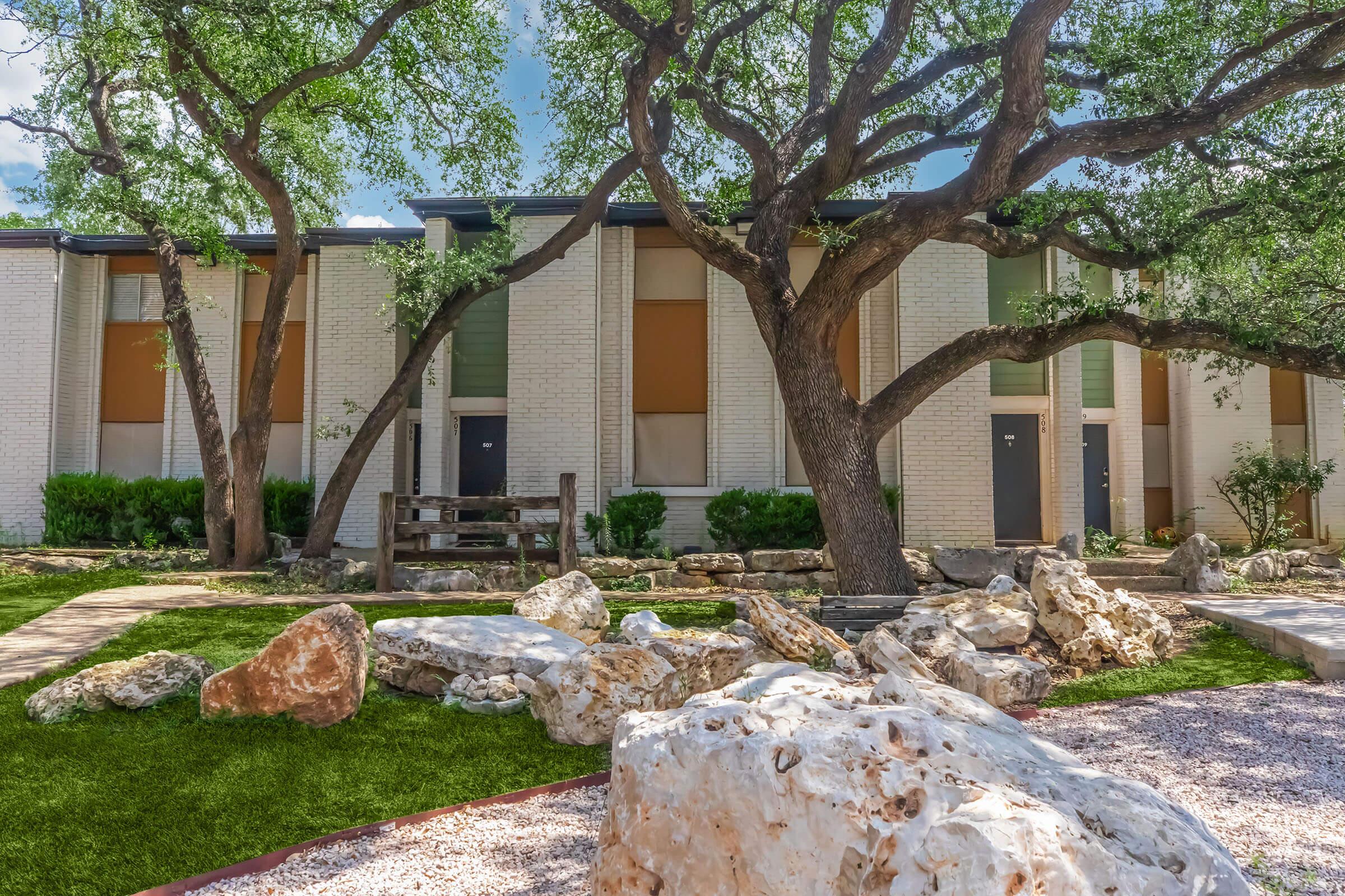 A view of an apartment complex featuring two-story buildings with a mix of green and orange shades on the windows. In the foreground, large rocks and a landscaped area with artificial grass and pathways are visible, along with a prominent tree providing shade.