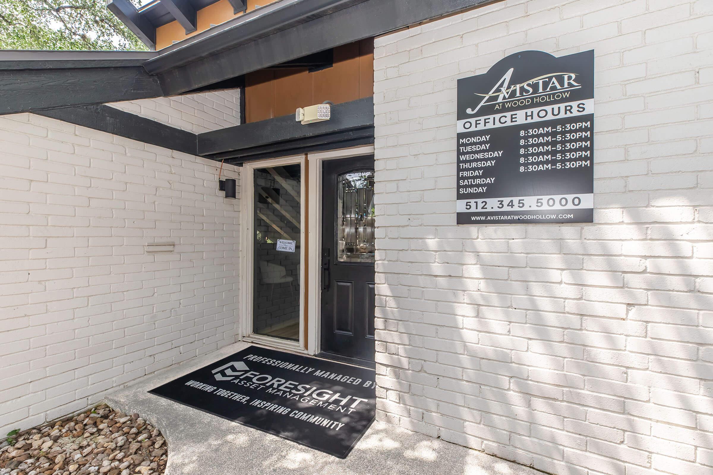 Entrance of a brick building featuring a door with glass panels. A sign on the wall lists office hours for each day of the week, and a welcome mat is placed at the entrance. The building is associated with Foresight Property Management.