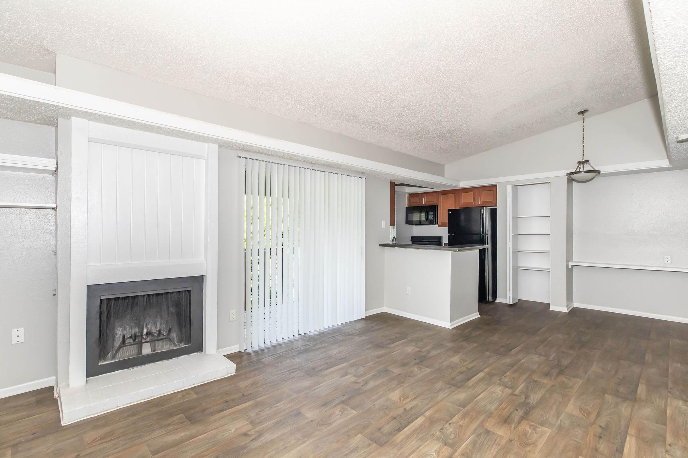 A spacious living area featuring a fireplace, large windows with vertical blinds, and an open kitchen with wood cabinetry. The flooring is a light wood look. The kitchen includes black appliances and a pantry, creating a bright and inviting atmosphere.
