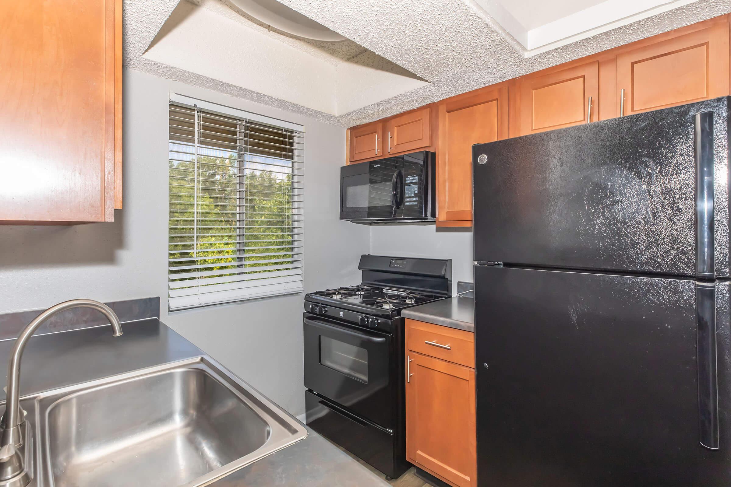 A modern kitchen featuring wooden cabinets, a black refrigerator, a gas stove, and a microwave. There is a stainless steel sink and a window with blinds allowing natural light to enter. The countertop is gray, and the kitchen has a clean, functional layout.