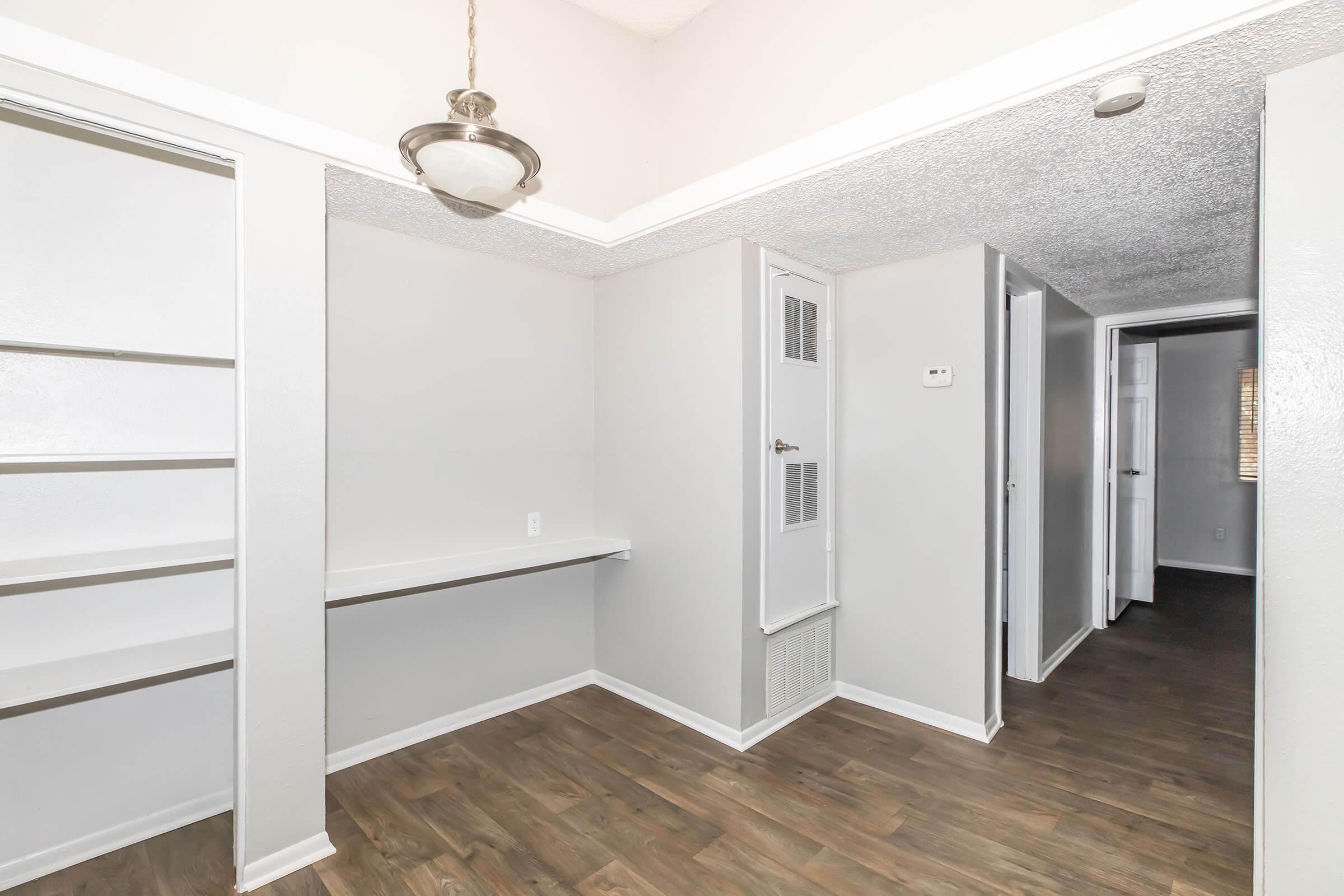 Interior view of a light-colored room featuring a small built-in shelf, a wall-mounted light fixture, and a doorway leading to another room. The floor is covered in dark laminate wood, adding warmth to the space. The walls are painted in neutral tones, complementing the overall design.
