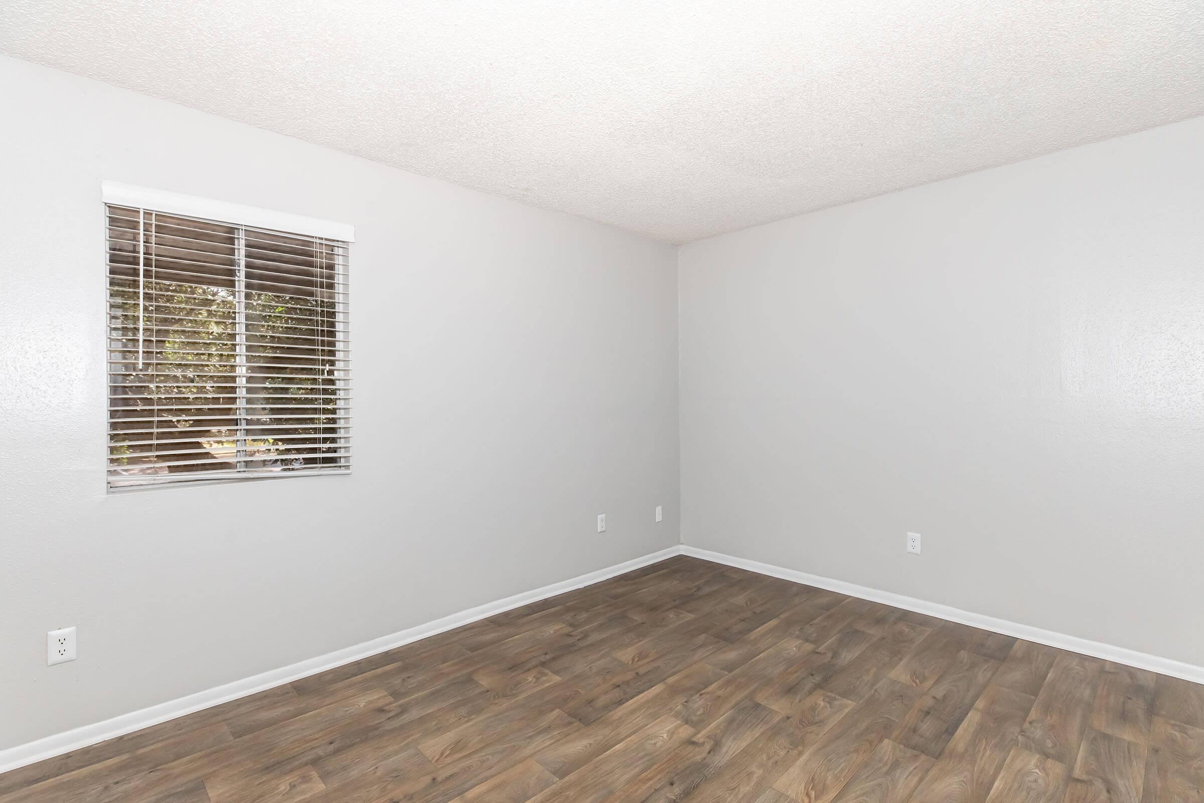 Empty room with light gray walls, a window with white blinds, and a wooden floor. The space is well-lit and has no furniture, creating an open and airy atmosphere.