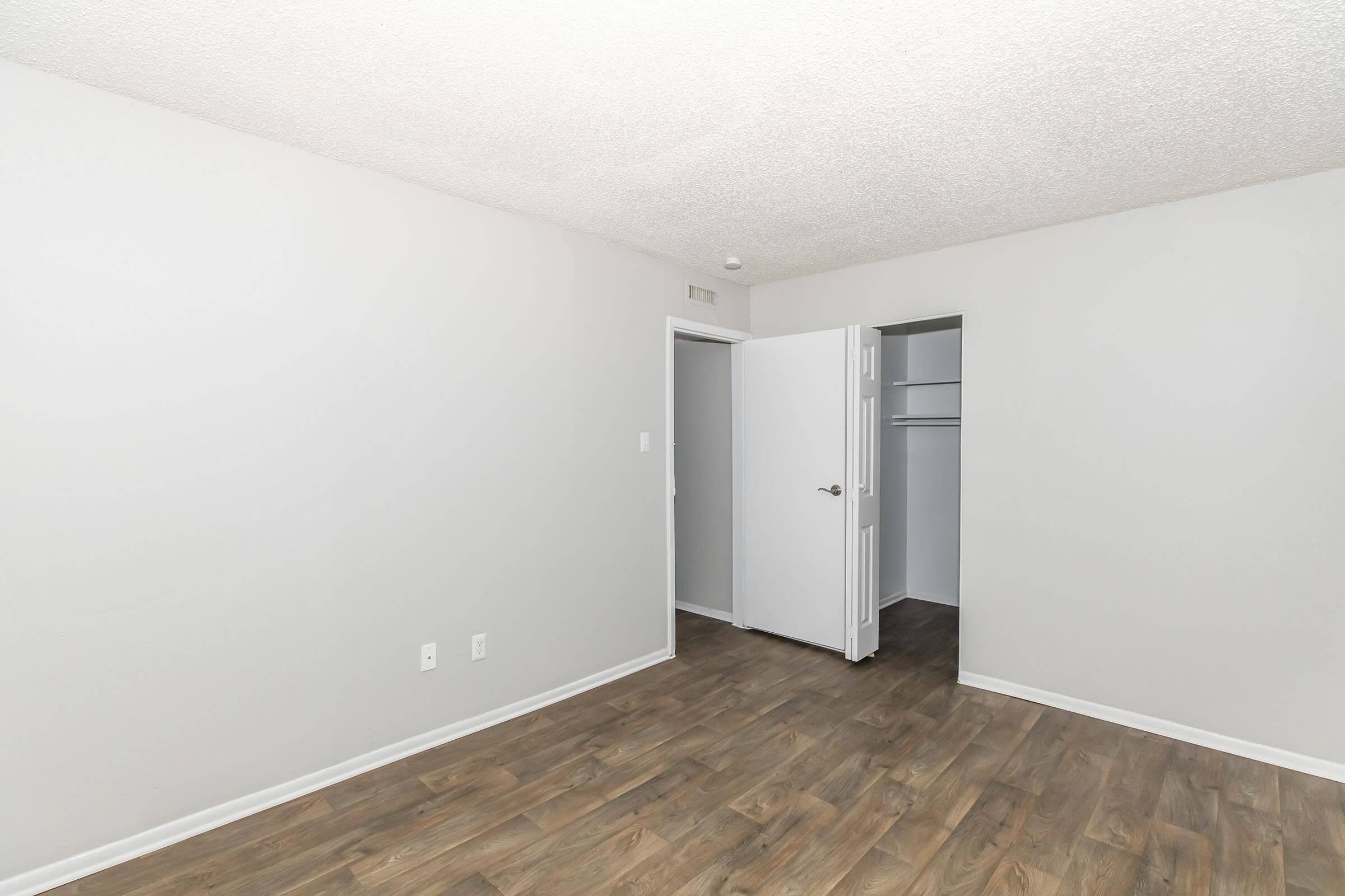 An empty room with light gray walls and a textured ceiling. There is a wooden floor and an open white door leading to a closet on the right. The room has a spacious feel, suitable for various furniture arrangements. No decorations or furnishings are present.
