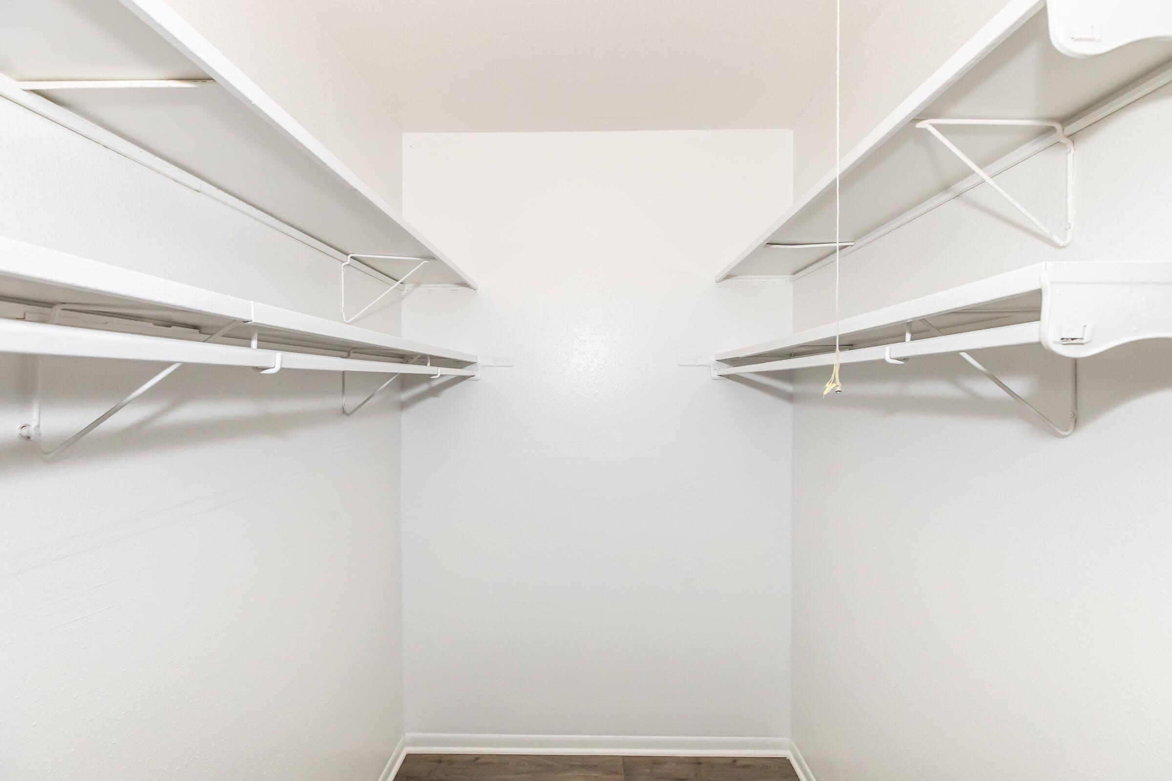 A clean, empty closet with white walls and metal shelving on both sides. The wooden floor is visible, and there is a light cord hanging from the ceiling, but no items or decorations are present inside the closet. The overall appearance is minimalistic and organized.