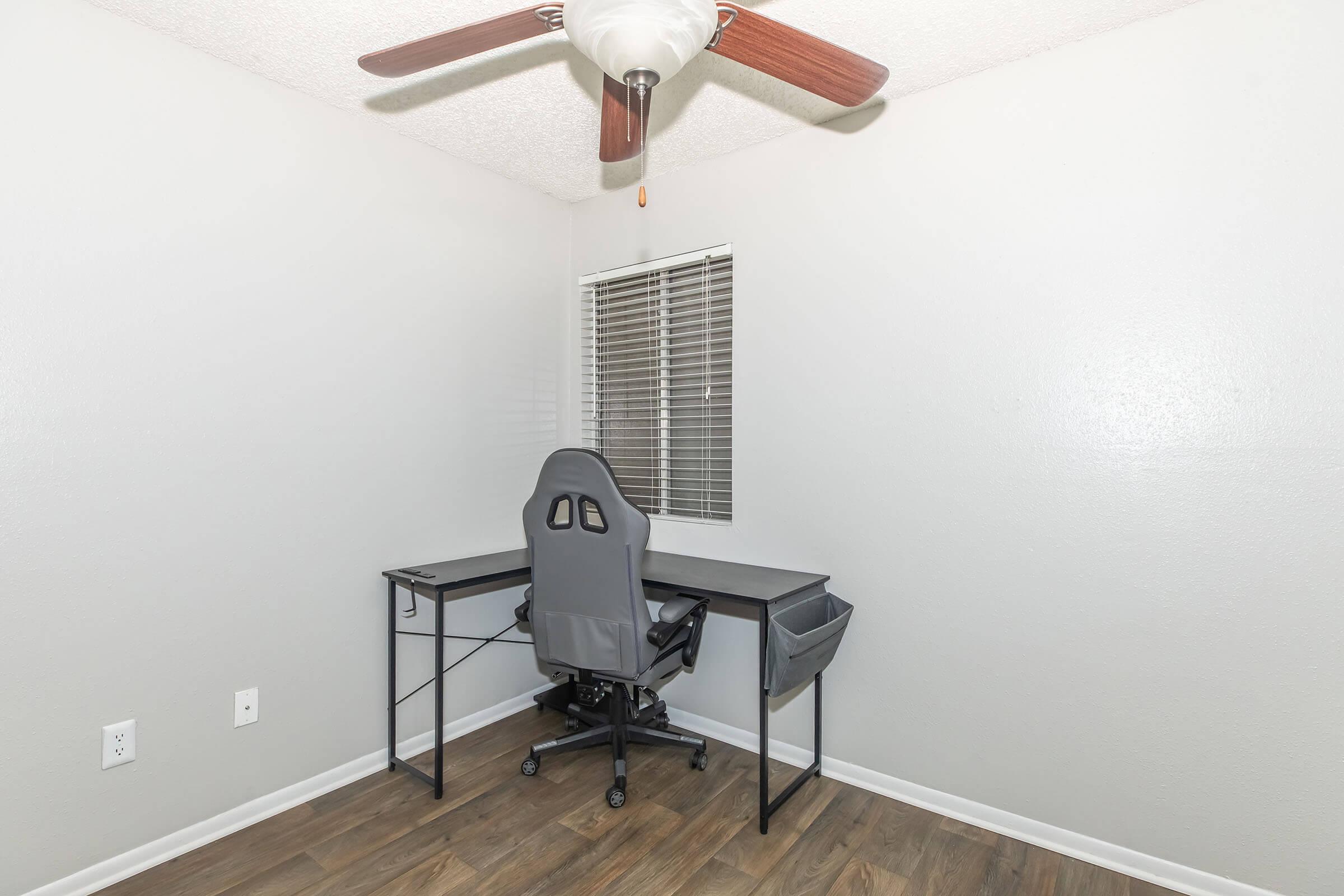 A minimalist home office setup featuring a black desk and a gray office chair. The room has light gray walls, a ceiling fan with wooden blades, and a window with blinds providing natural light. The floor is wooden, contributing to a clean and modern aesthetic.