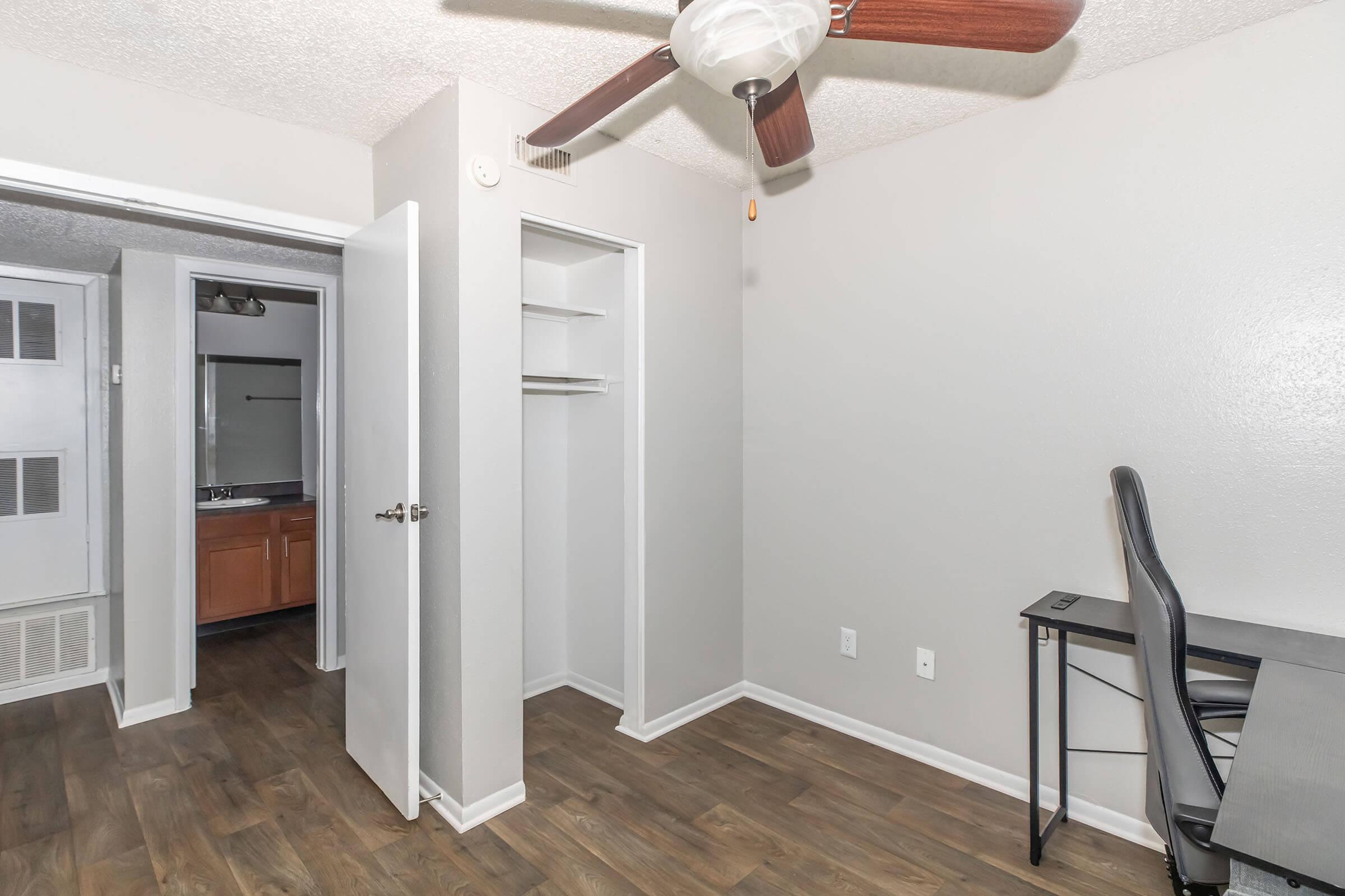 Room interior featuring a ceiling fan, a small desk, and a closet with open sliding doors. The walls are painted light gray, and the floor is hardwood. A doorway leads to another area of the space, suggesting an open layout.