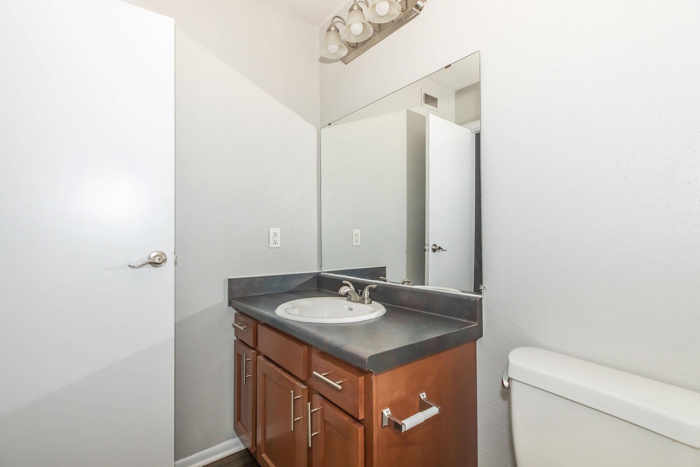 A bathroom with a gray countertop and wooden cabinetry. There is a sink with a mirror above it, a toilet on the right, and a closed white door in the foreground. A second door is partially open in the background, leading to a light-colored hallway. The walls are painted in a light shade.
