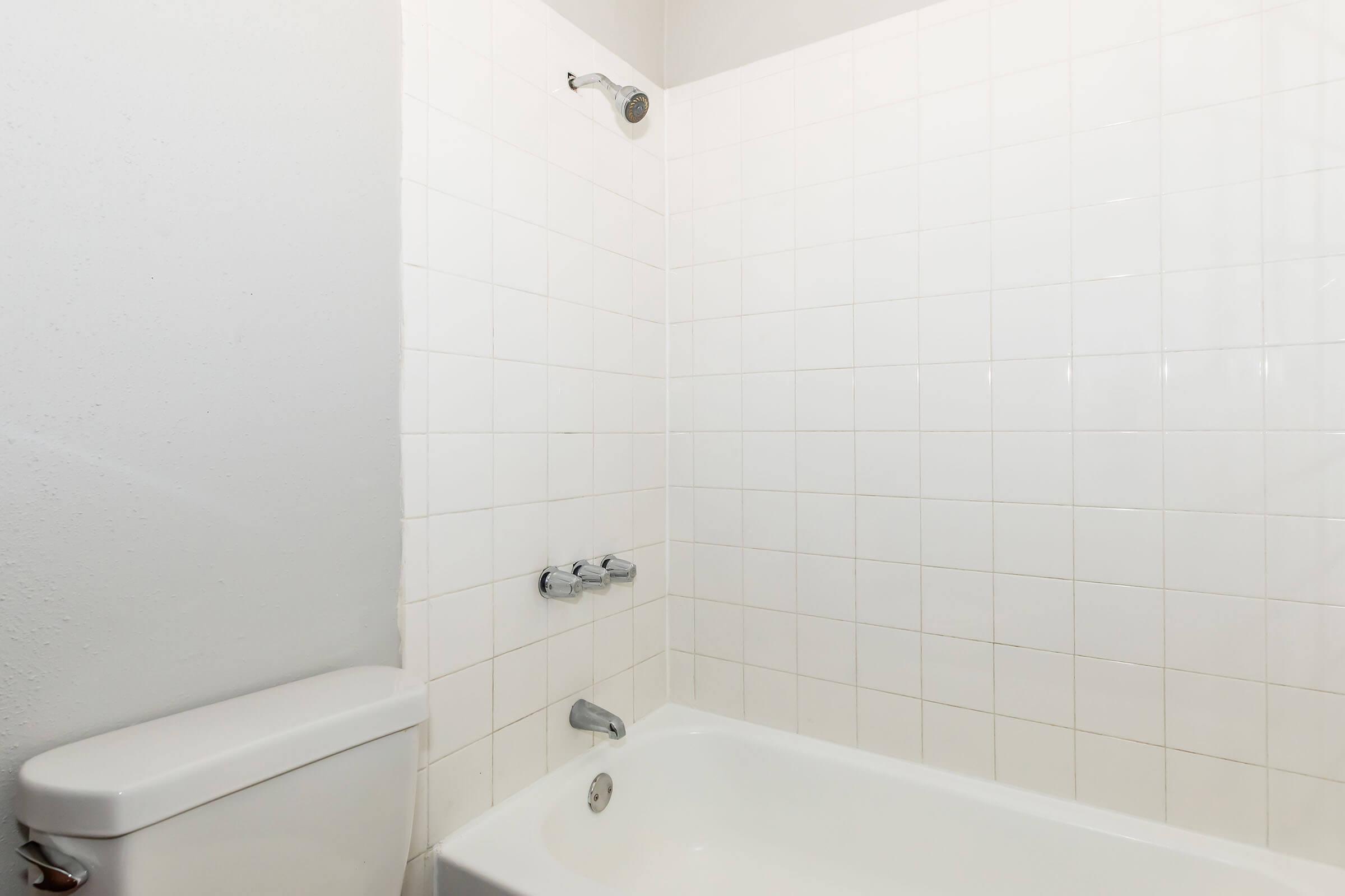 A clean bathroom featuring a white tiled wall with a bathtub. The bathtub is accompanied by a showerhead mounted on the wall, and a toilet is visible beside the tub. The overall aesthetic is simple and minimalistic, with a focus on functionality.
