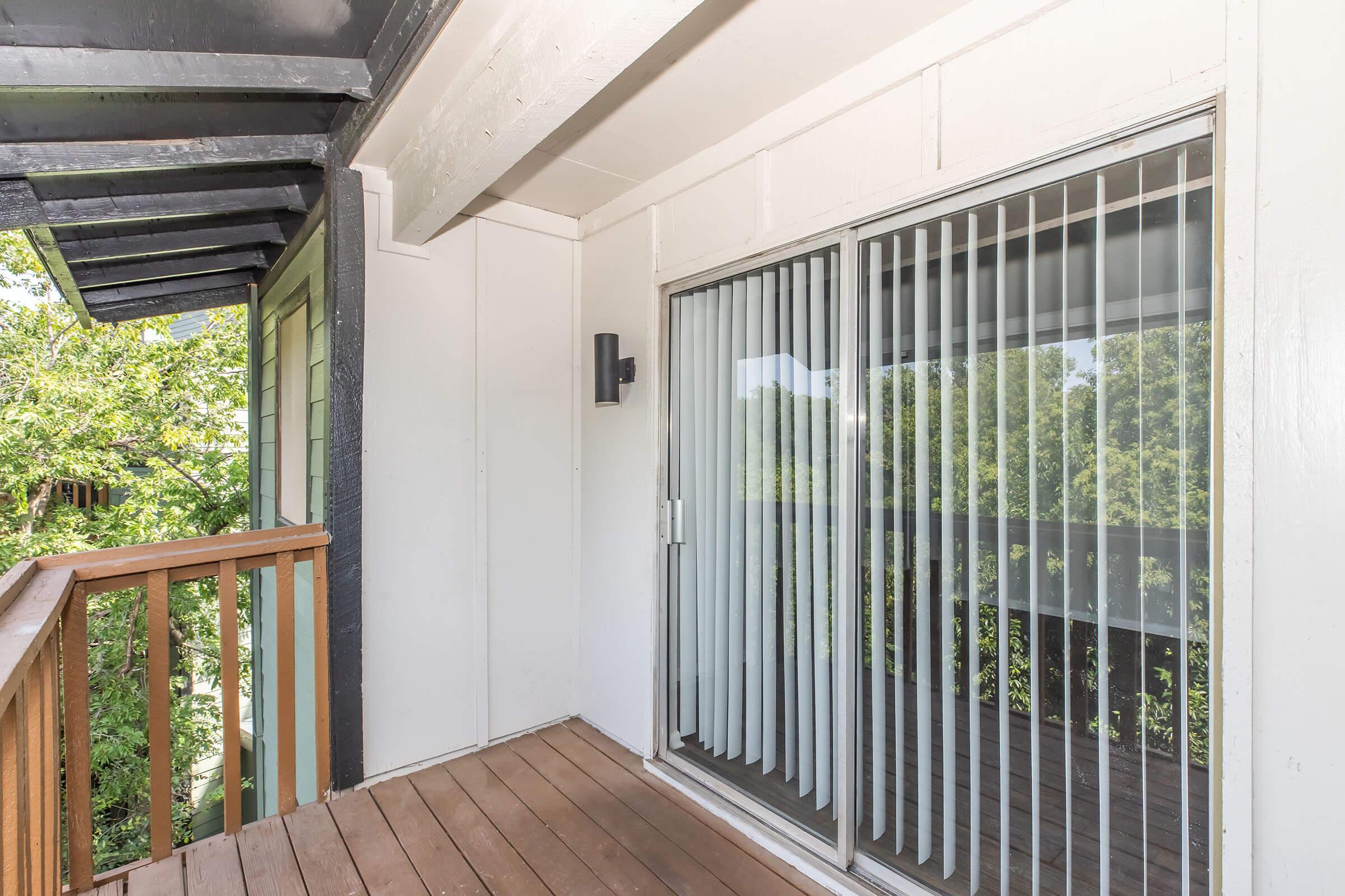 A wooden balcony with a railing, featuring sliding glass doors that have vertical blinds. The walls are painted white, and there is a small exterior light fixture near the doors. Green foliage is visible outside, suggesting a natural, peaceful setting.