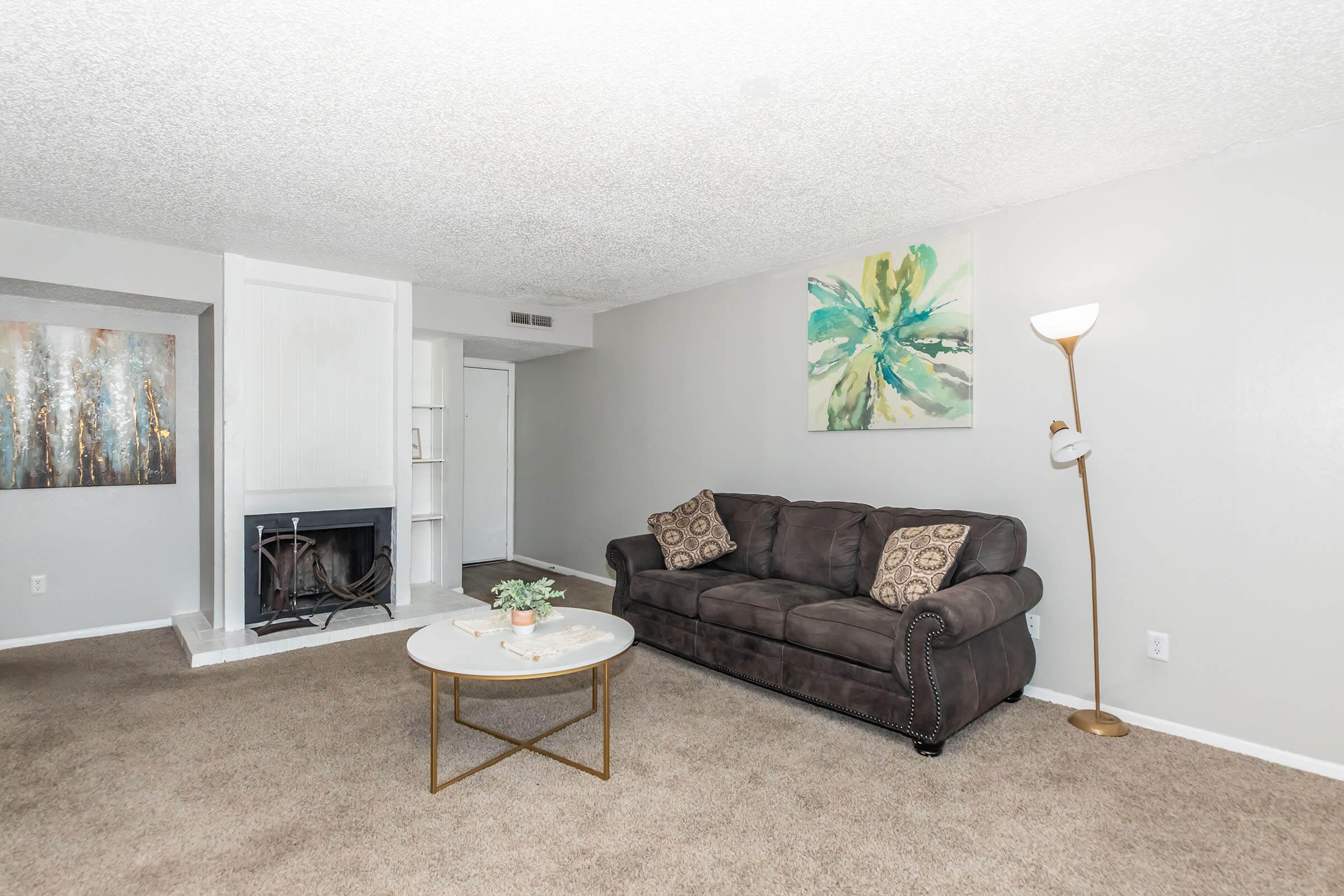 A cozy living room featuring a dark brown couch with decorative pillows, a round marble coffee table with a small plant on top, and a standing lamp. The walls are painted light gray, and there is a fireplace on one side. A colorful abstract artwork is hung on the wall, adding a vibrant touch to the space.