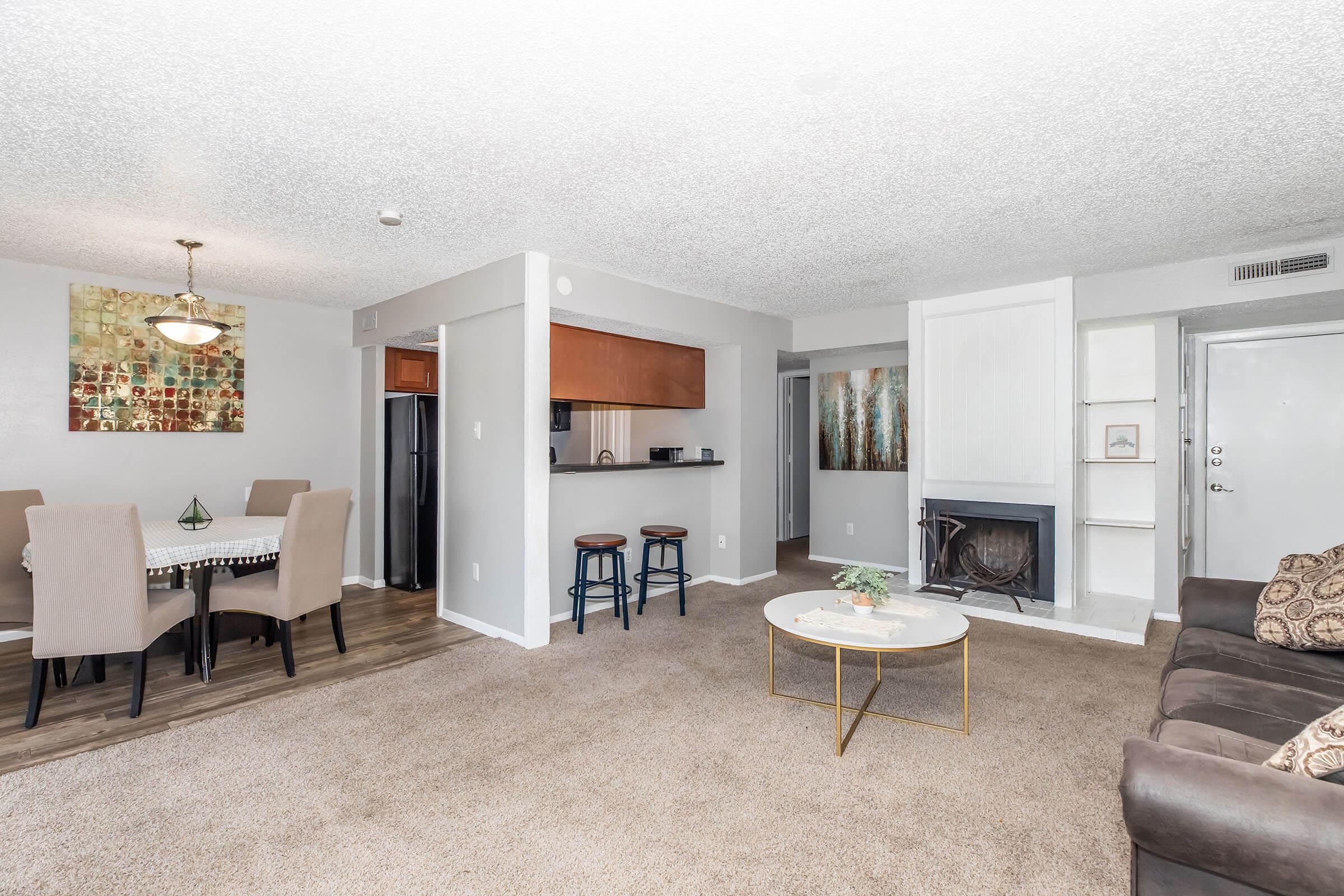 A spacious living area with a dining table, bar stools at a kitchen counter, and a cozy fireplace. The decor features a light-colored wall with artwork, plush carpeting, and contemporary furniture, creating a welcoming atmosphere. Natural light brightens the space, with a view of the entrance.