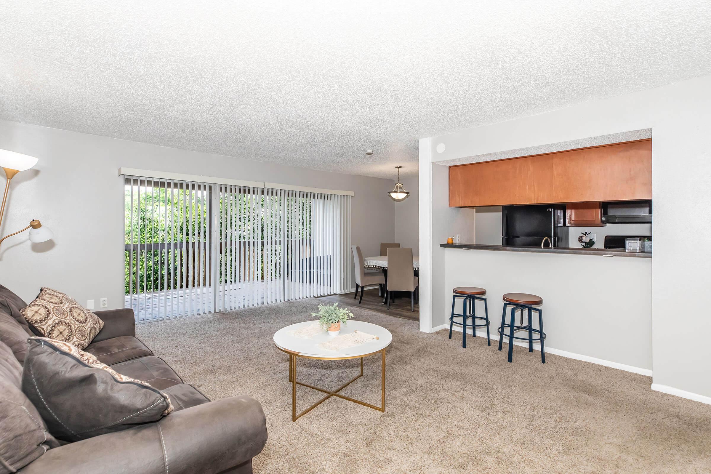 A cozy living room with a brown sofa and decorative pillows, a round coffee table with a small plant, and two barstools. The space features a sliding glass door with vertical blinds, leading to a balcony. A dining area with a table and a light fixture is visible, with a kitchen area in the background.