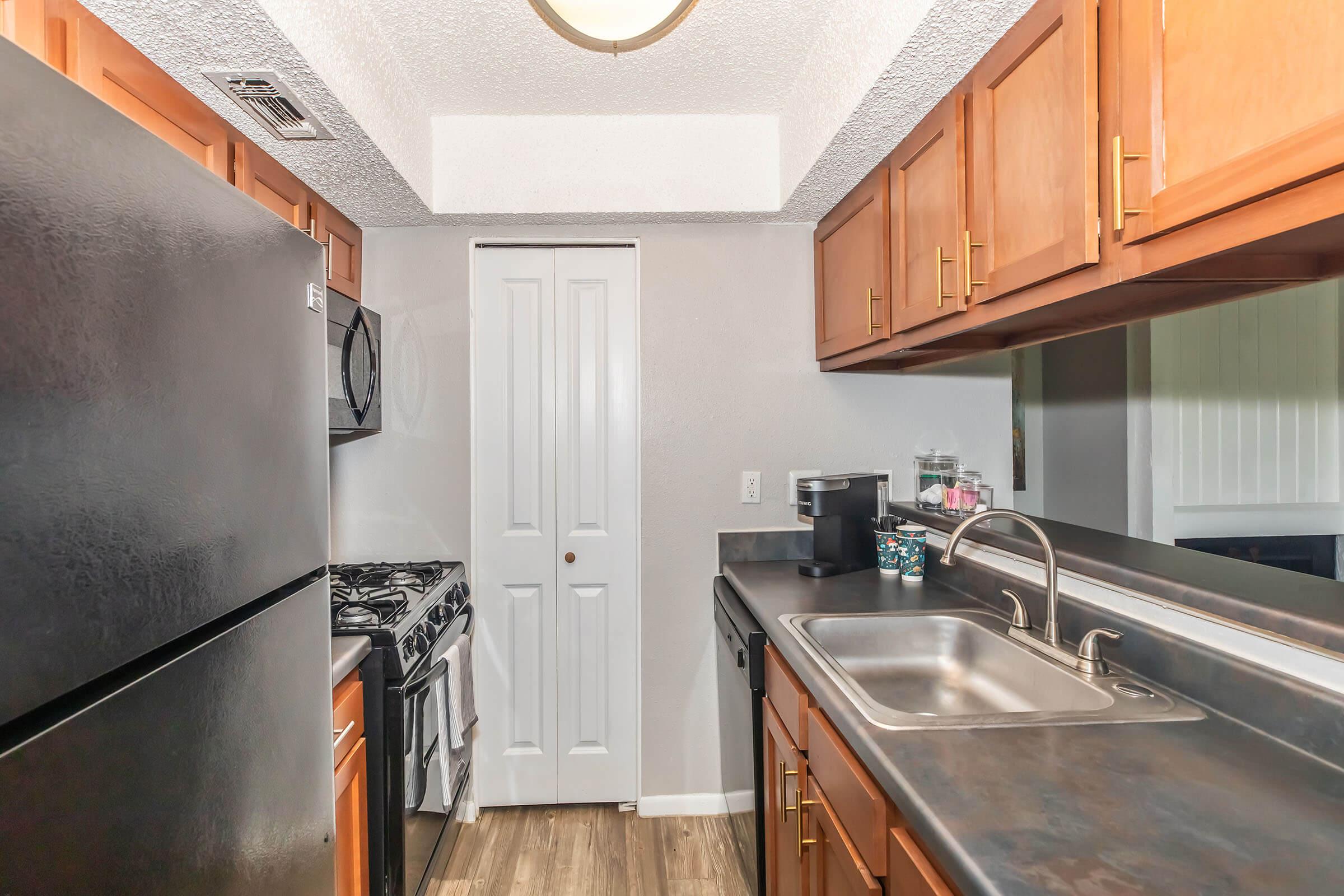 View of a modern kitchen featuring dark wood cabinetry, stainless steel appliances including a gas stove and refrigerator, a double sink, and a doorway leading to another room. The countertop is dark, and light wood-like flooring complements the space. The overall atmosphere is clean and well-organized.