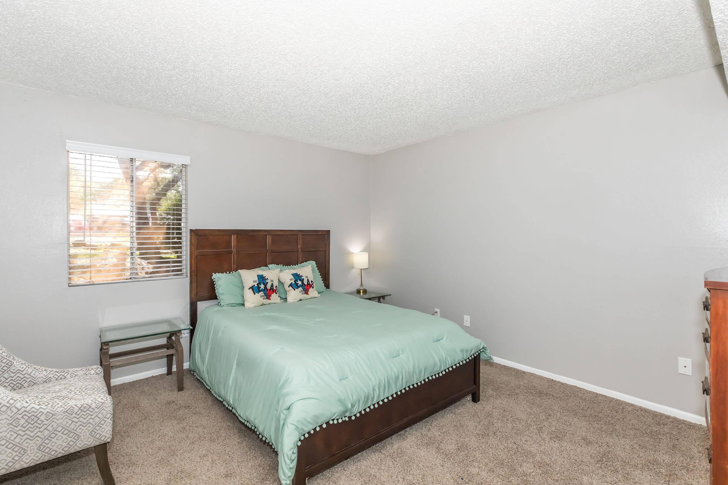 Cozy bedroom featuring a queen-size bed with a mint green comforter and decorative pillows. A small bedside lamp and a chair are present, with a window providing natural light. The walls are painted in a light gray, and the carpeted floor adds warmth to the space. 