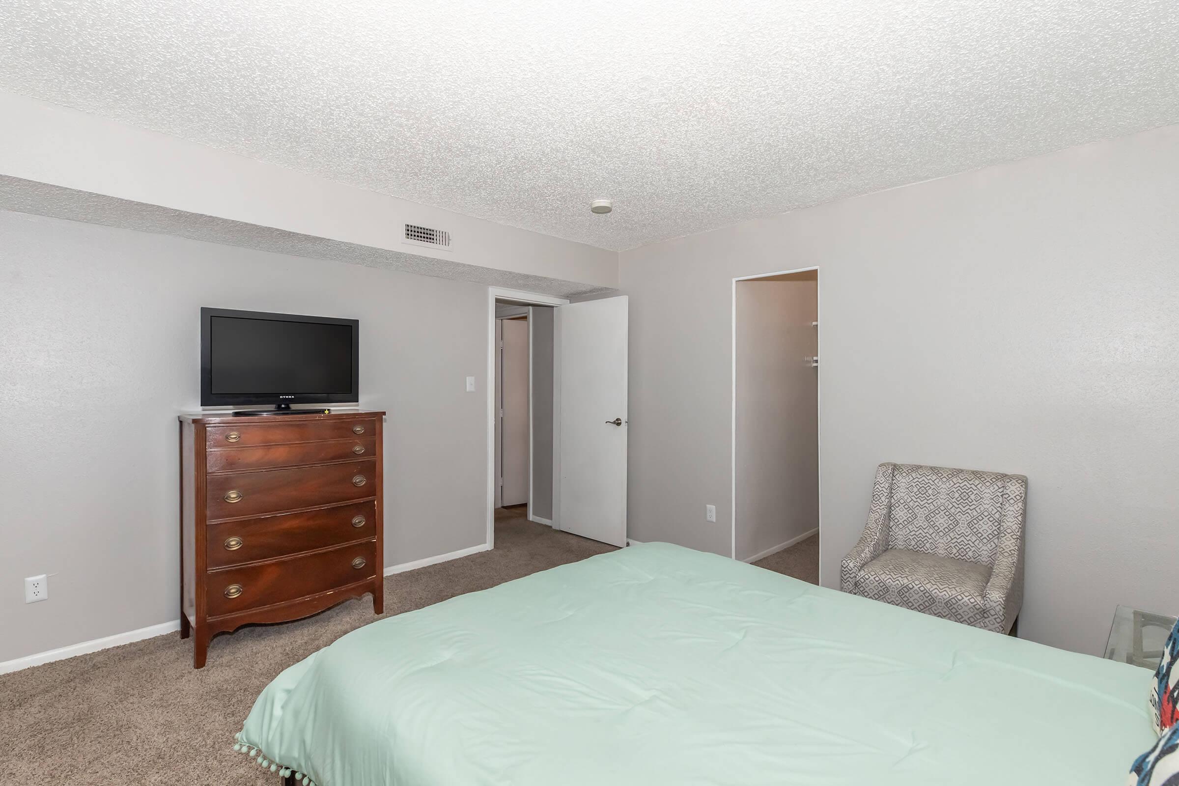 A spacious bedroom featuring a light green bedspread, a wooden dresser with a television on top, a patterned armchair, and a door leading to a closet. The walls are painted in neutral tones, and the carpeted floor adds warmth to the space.