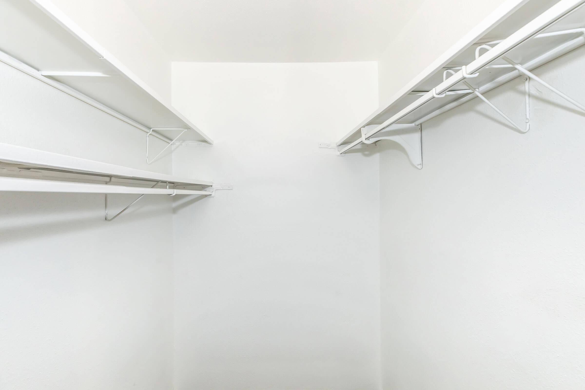 Empty walk-in closet with white walls and white wire shelves. The shelves are positioned on either side, creating a minimalist and spacious appearance. Natural light might be illuminating the space, emphasizing its simplicity and cleanliness.