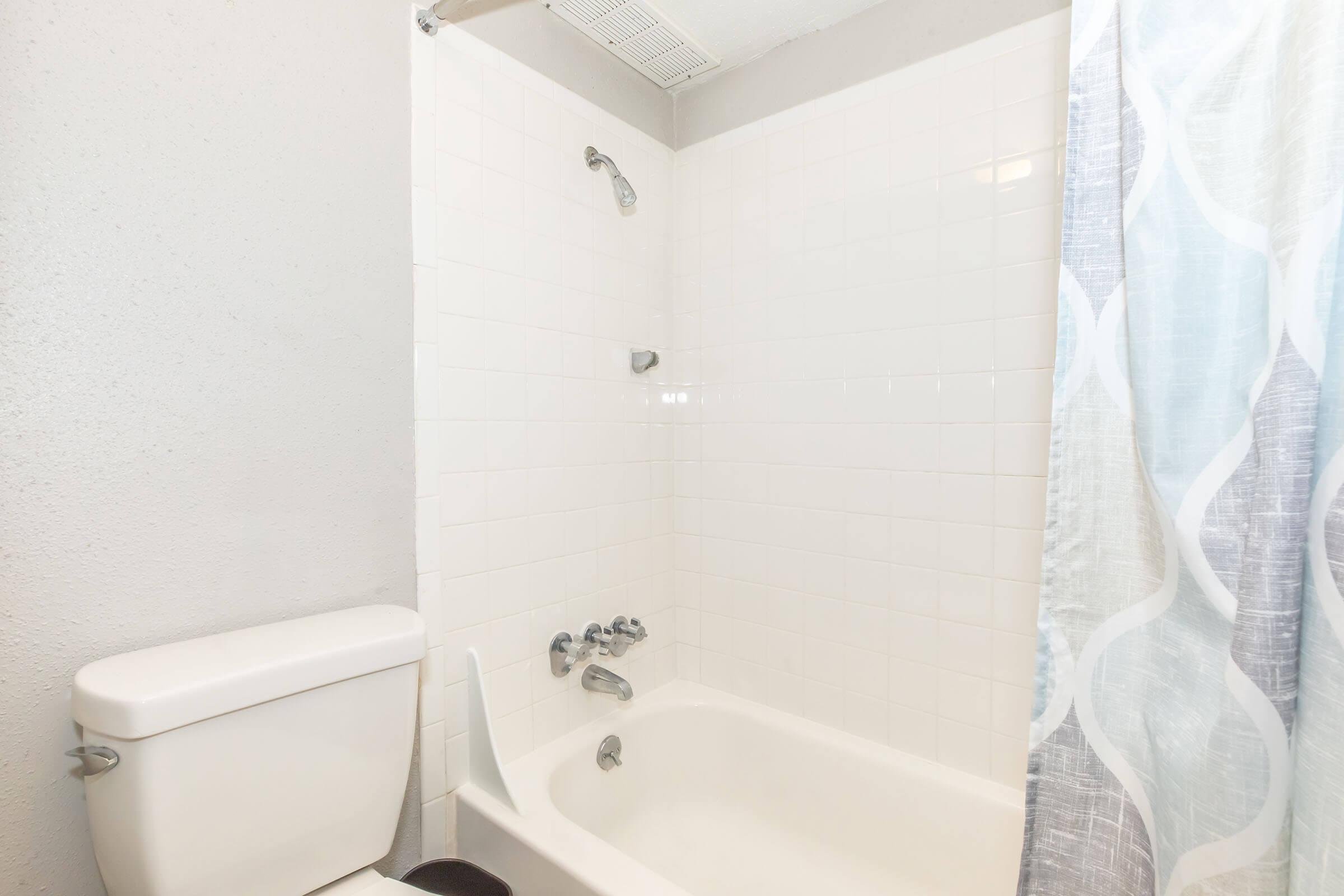 A clean bathroom featuring a shower stall with a clear shower curtain, a white bathtub, and a toilet. The walls are light-colored tiles, and there is a wall-mounted showerhead. The shower curtain has a subtle pattern, and the overall space is well-lit and tidy.