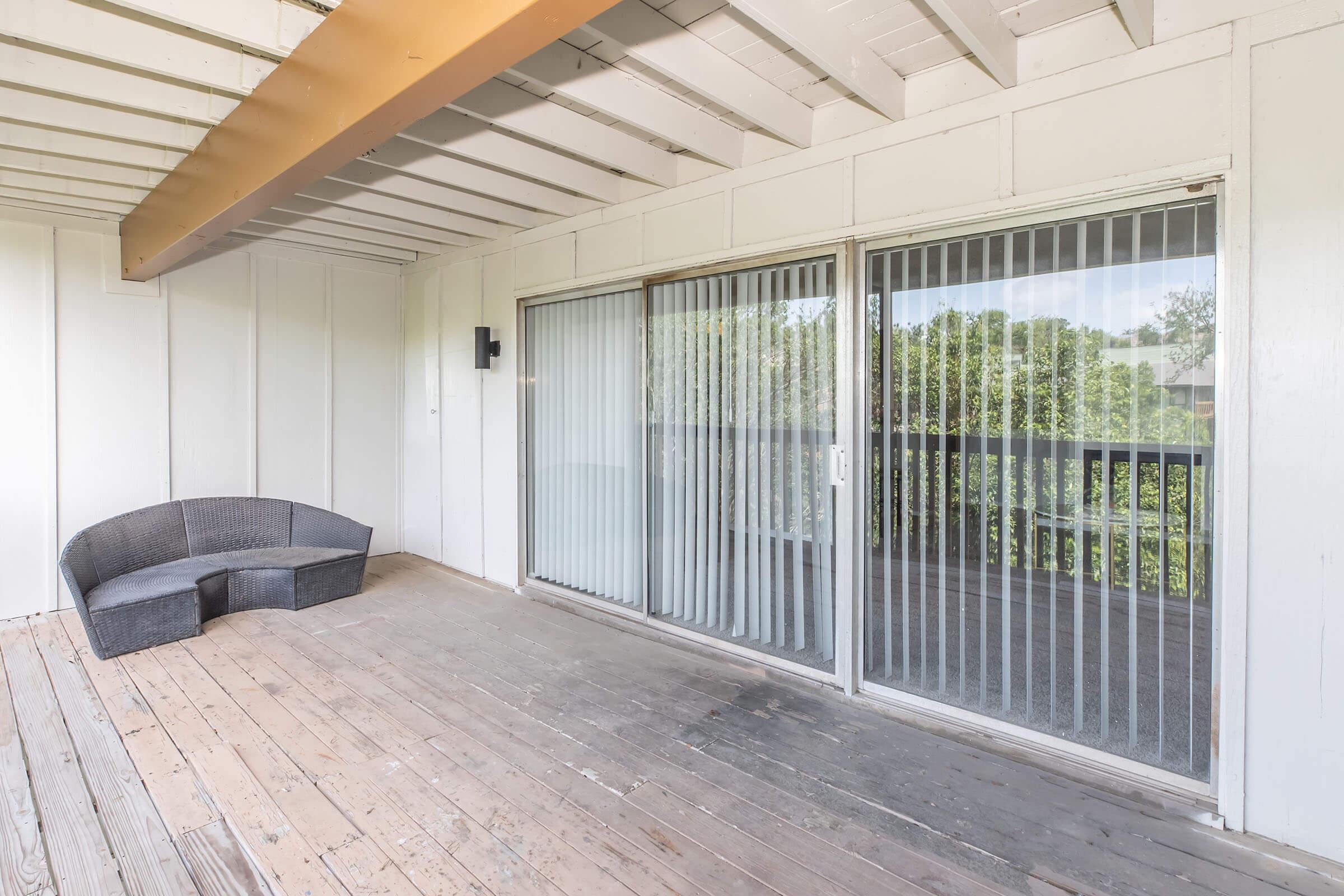 A spacious, empty outdoor balcony featuring a curved gray wicker seating area. Large sliding glass doors with vertical blinds lead to an interior room, and the wooden floor is unfinished. The surrounding area is green, visible through the glass.