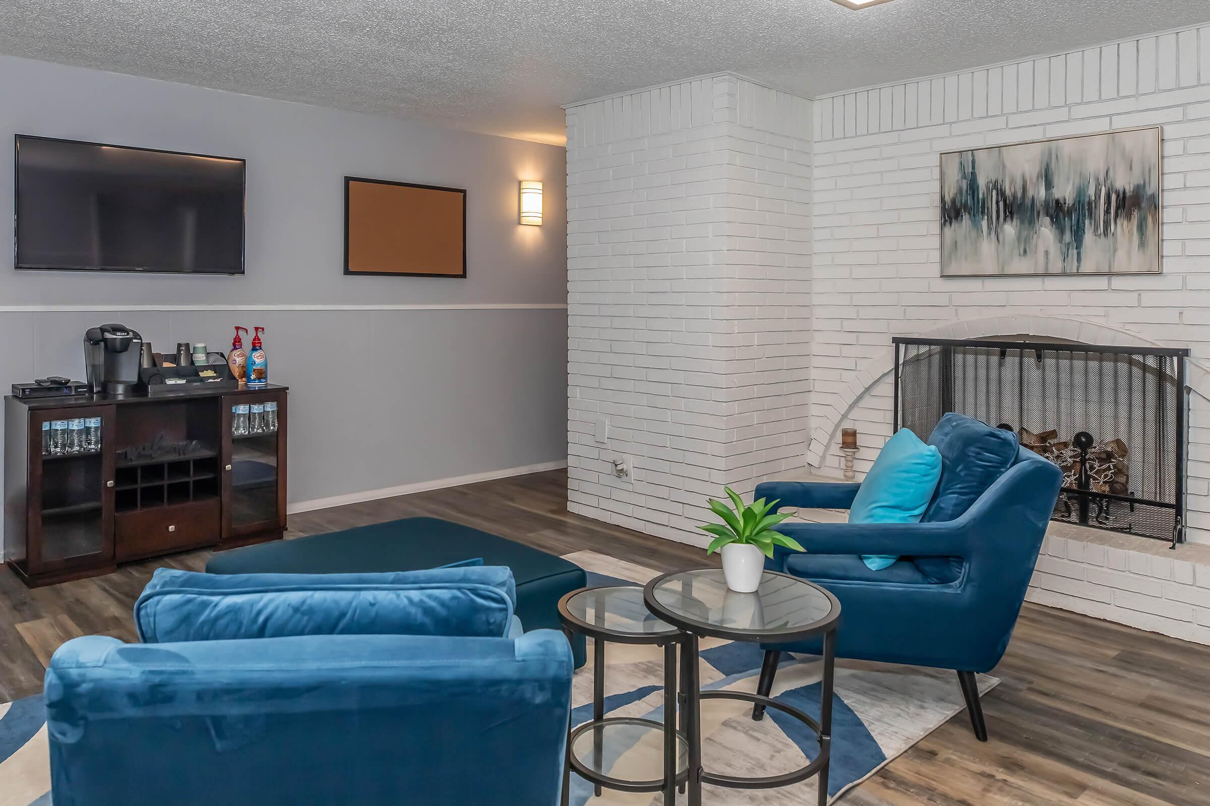 A cozy living room featuring two teal chairs, a small round table, and a modern fireplace. The walls are painted in light gray and white, with a flat-screen TV mounted on one wall. There is a decorative painting above the fireplace and a coffee station in the corner. Natural light enhances the inviting atmosphere.