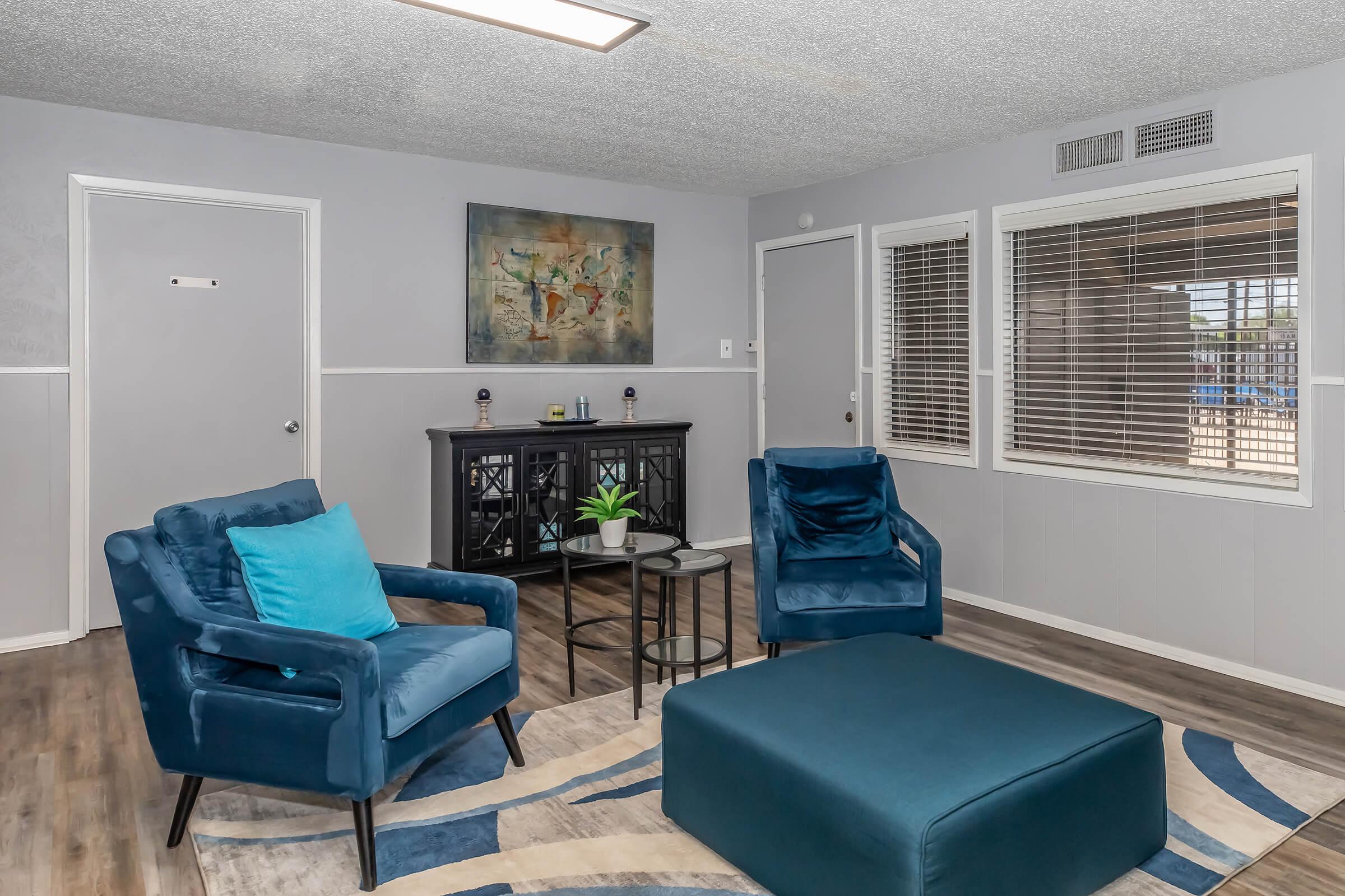 A cozy living room featuring two blue armchairs with a teal pillow, a small round table, and a large ottoman. A black cabinet holds decorative items, and a piece of abstract art hangs on the wall. Natural light enters from a window with blinds, enhancing the warm, inviting atmosphere.
