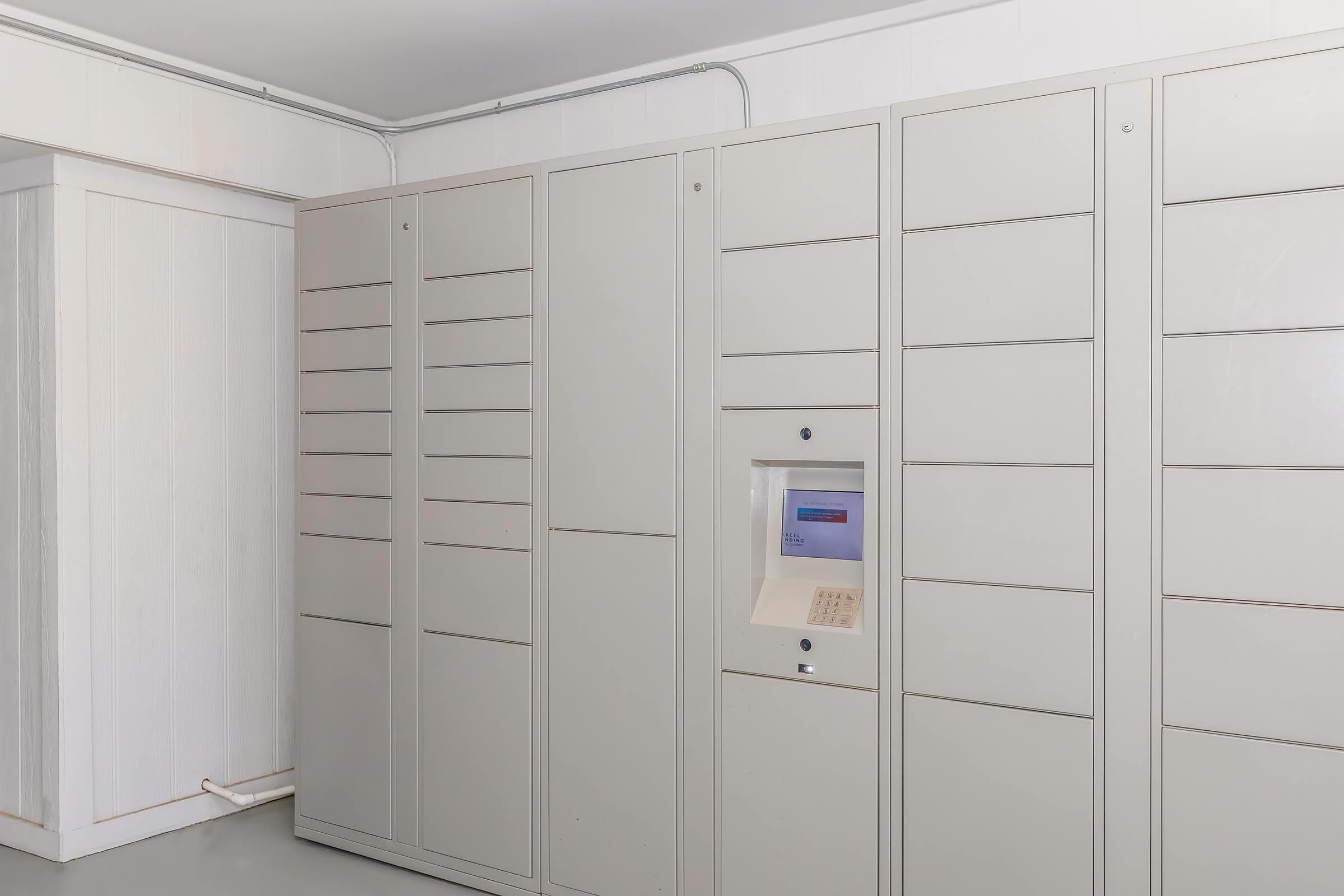 A row of light gray lockers lining a wall in a modern interior setting. One locker features a digital panel in the center, likely used for accessing packages or deliveries. The room has a clean and minimalistic aesthetic, with a simple white wall adjacent to the lockers.