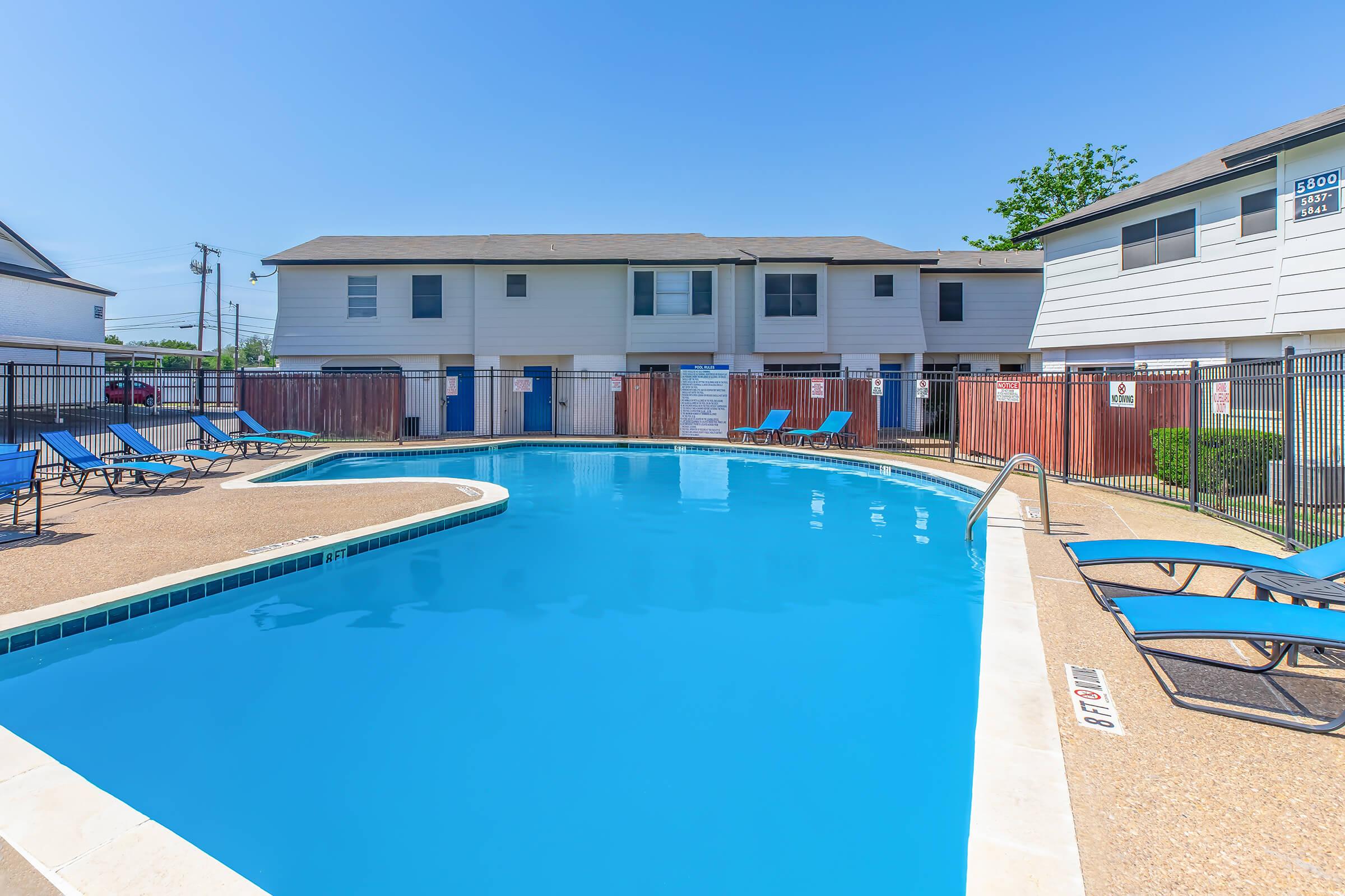 A clear blue swimming pool surrounded by lounge chairs and a fenced area. In the background, there are two-story apartment buildings, and the sky is bright and sunny. The pool area is well-maintained, inviting relaxation and leisure.