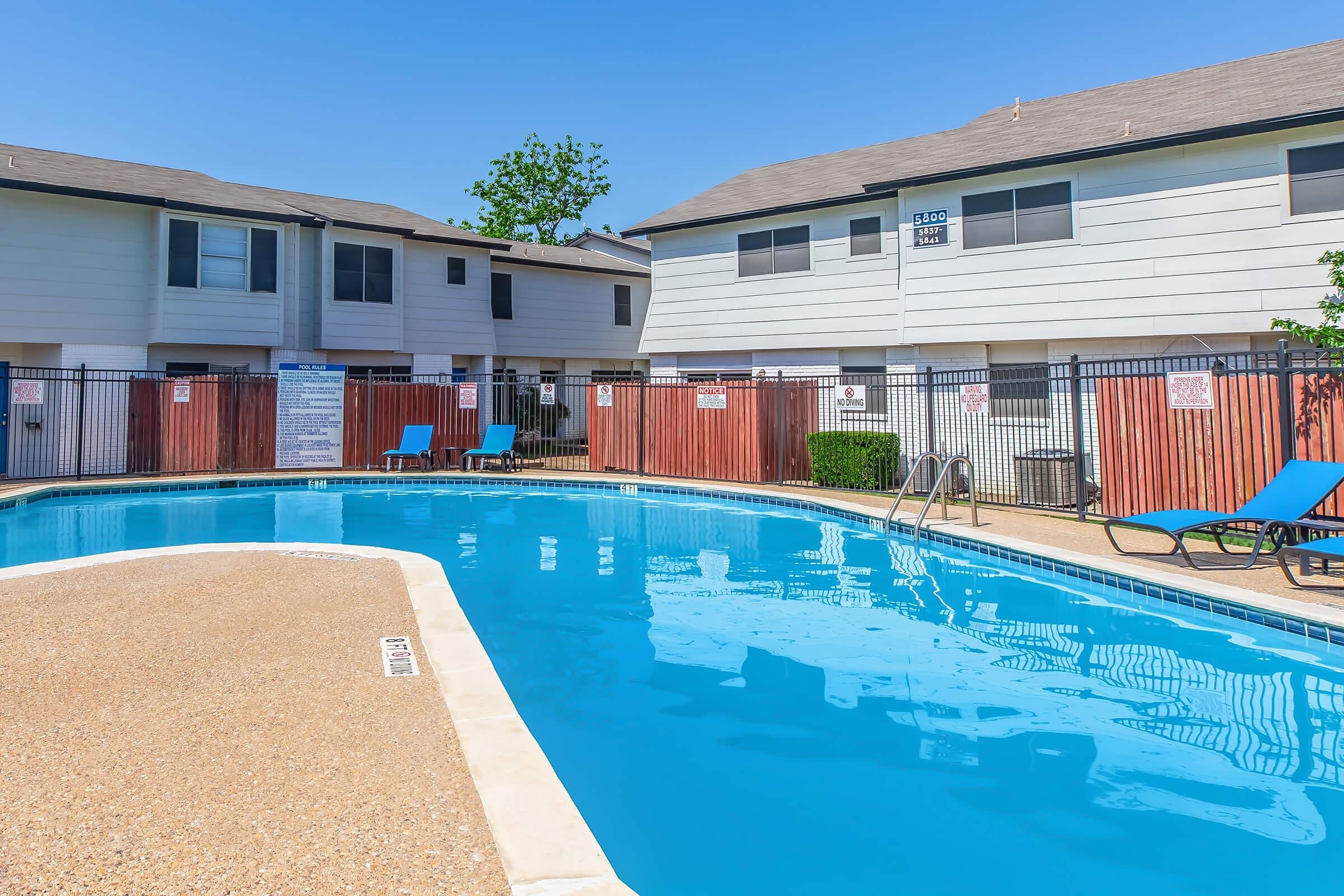 A clear blue swimming pool surrounded by lounge chairs and a fenced patio area. Apartment buildings are visible in the background under a bright blue sky. The scene is set in a well-maintained residential complex, creating a relaxing atmosphere.
