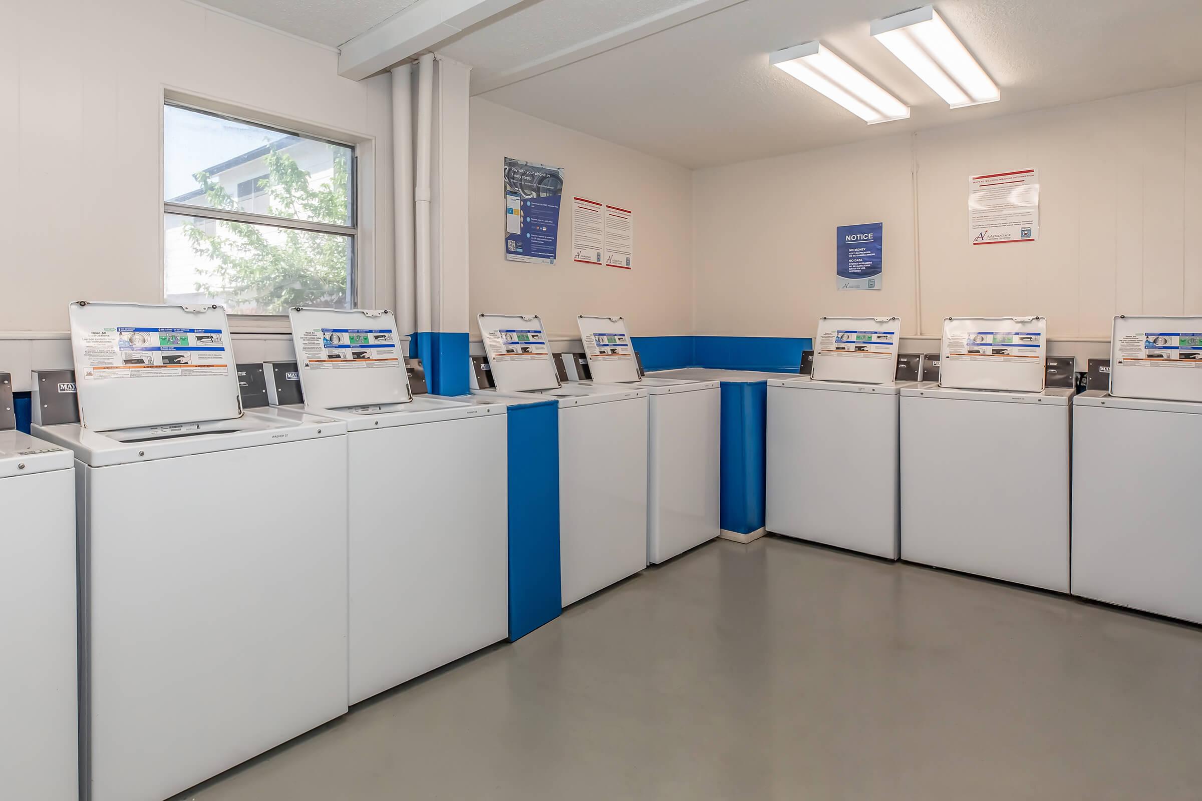 A clean and organized laundry room featuring multiple washing machines lined up against the walls. The space is well-lit with overhead lighting, and there are informational posters on the walls. The floor is gray, and the walls are painted white with blue accents.