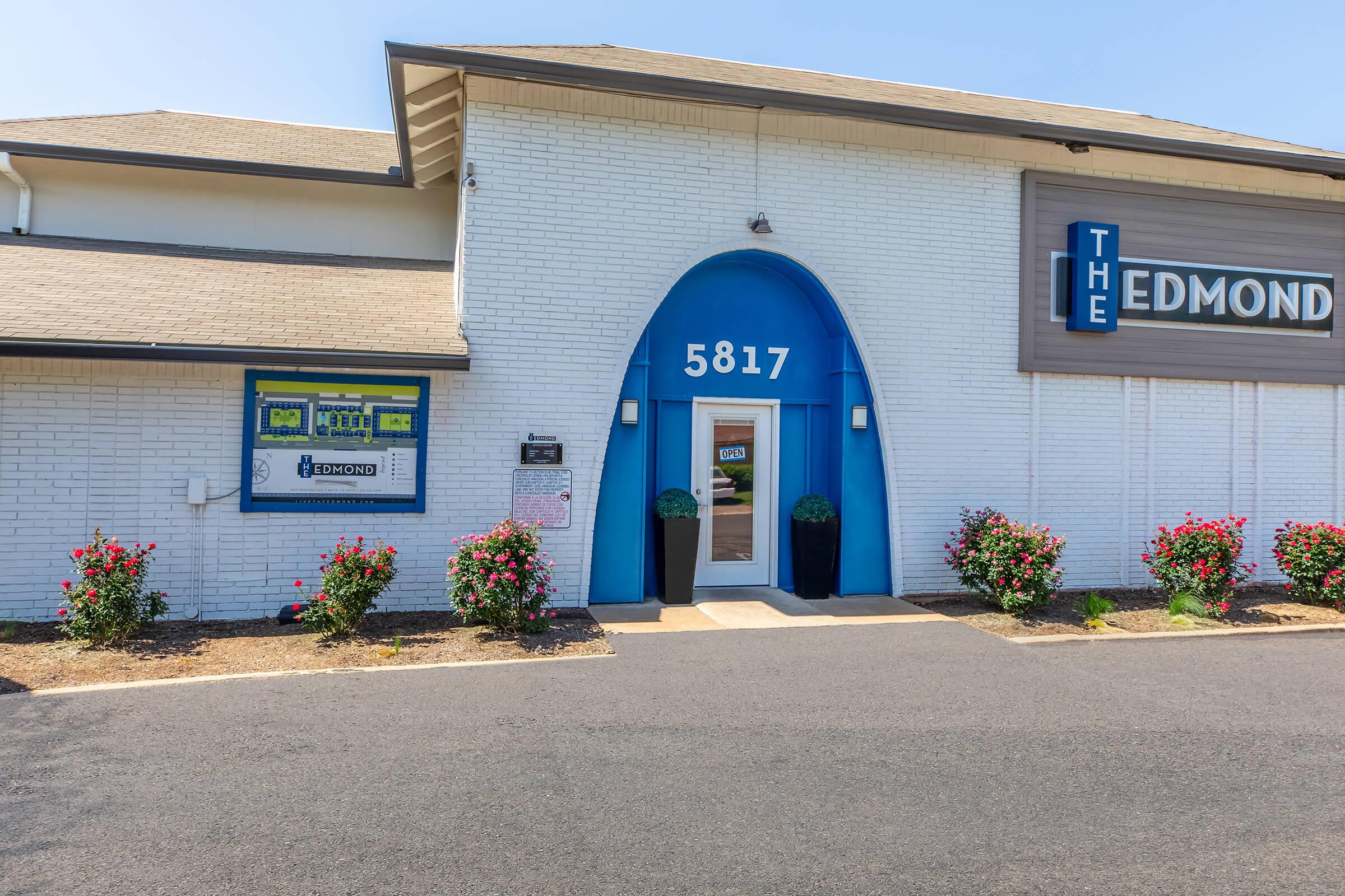 Exterior view of a white brick building featuring a blue entrance with the number 5817. Flower planters are placed on either side of the entrance, and rose bushes line the pathway. A sign displays the name "The Edmond" along with other signage prominently visible. Clear skies and a well-maintained landscape enhance the scene.