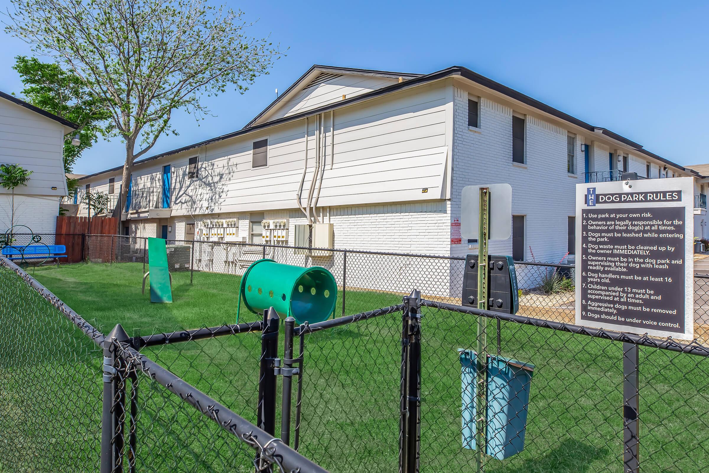 A fenced dog park area featuring green grass, a play structure, and a blue bench. A sign detailing dog park rules is displayed nearby. The background shows a white multi-unit building with windows and a tree. The scene is well-lit, indicating a sunny day.