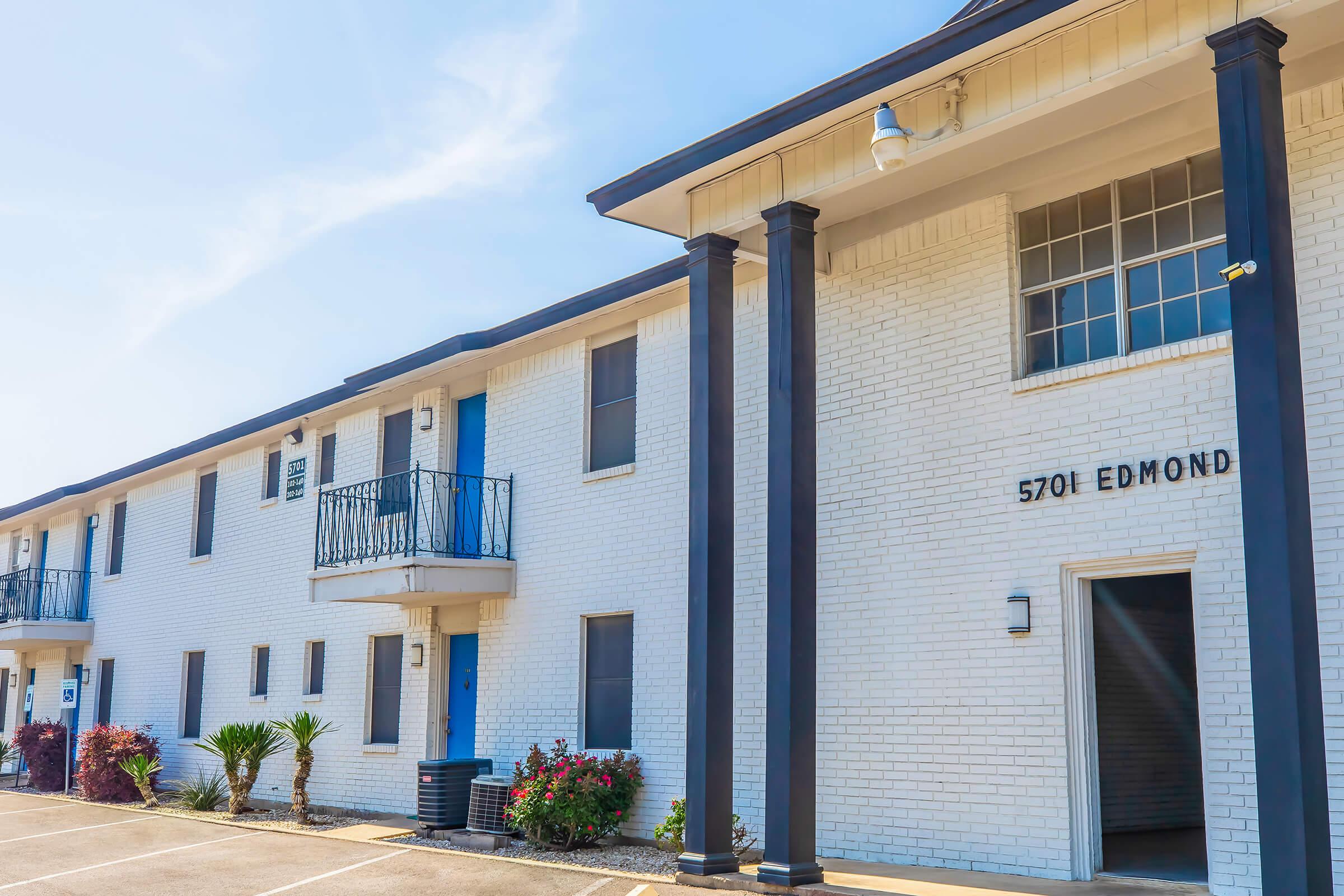 A brick apartment building with a blue door and balcony, featuring white walls and black columns. The building has multiple windows, some with shutters, and is surrounded by small plants and flowers. The sign indicates the address as "5701 EDMOND." Clear blue sky in the background.