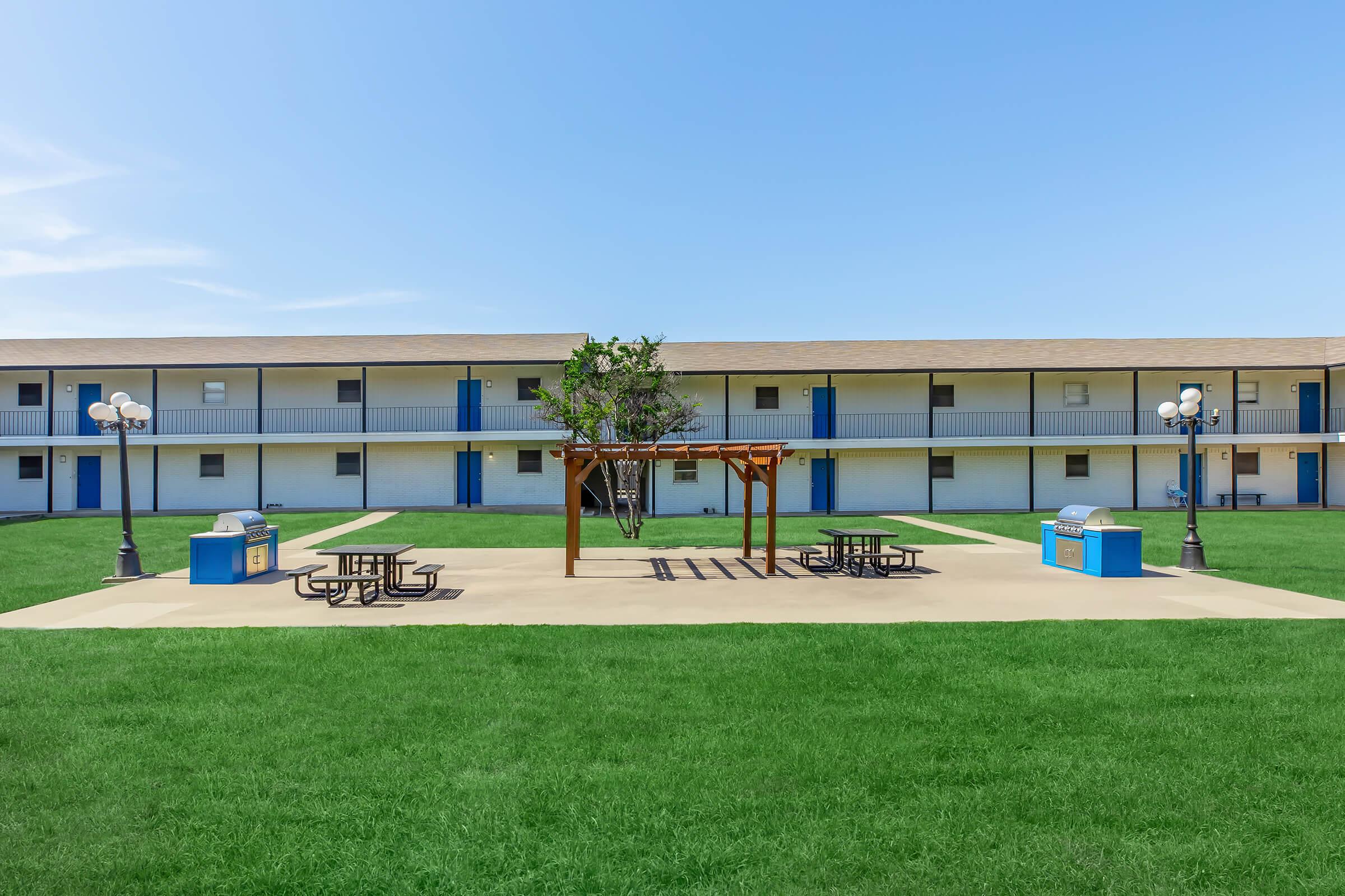 A courtyard area featuring a picnic table set under a wooden gazebo, surrounded by green grass. In the background, there are two-story buildings with blue doors and light-colored walls. Two trash bins are positioned near the picnic area. The sky is clear and blue.
