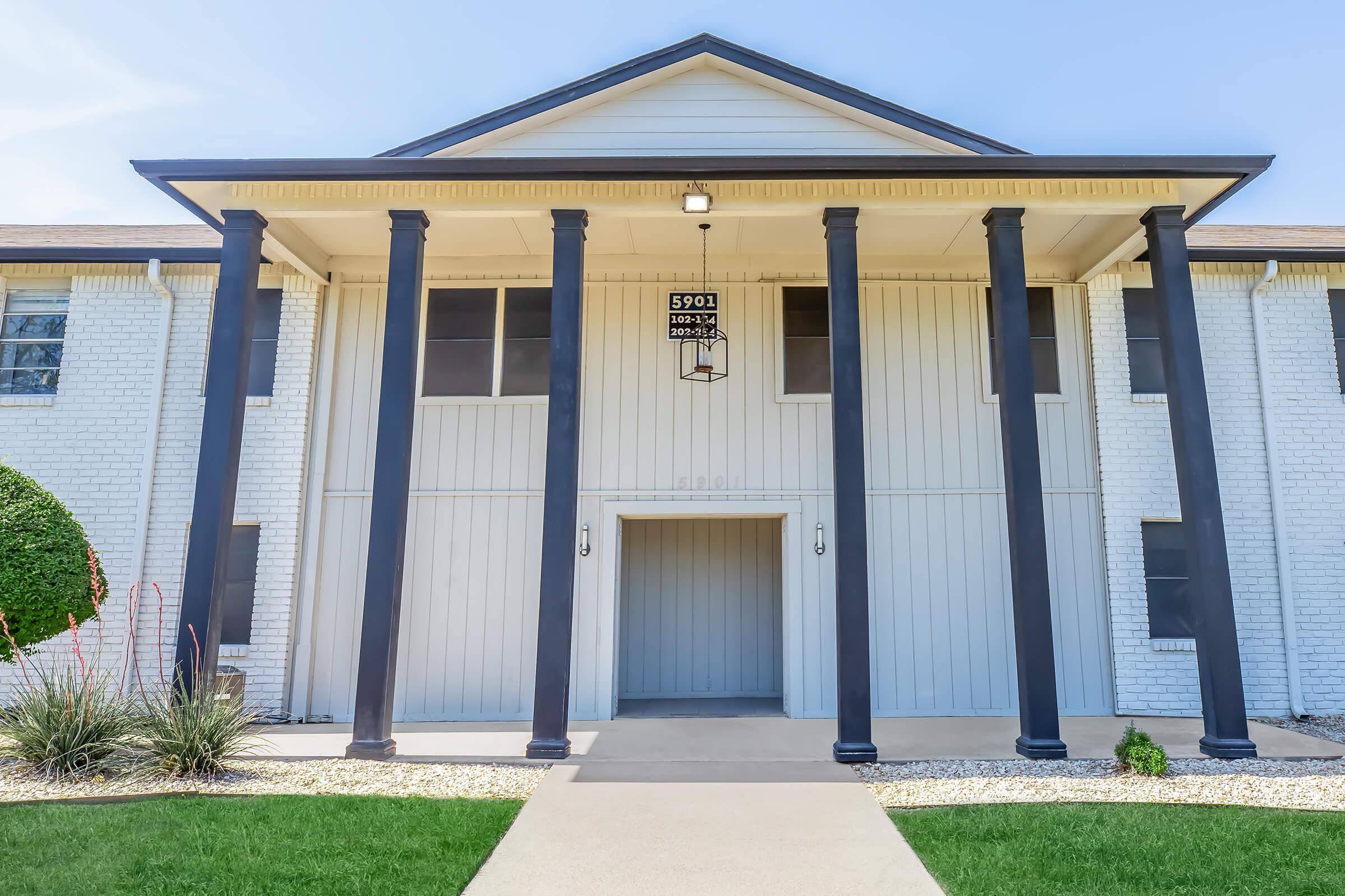 A large, modern building entrance featuring a tan façade with white brick accents. The entrance is framed by four tall black columns, and a sign hangs above the door. A pathway leads up to the entrance, surrounded by shrubs and decorative stones. Bright blue sky is visible above.